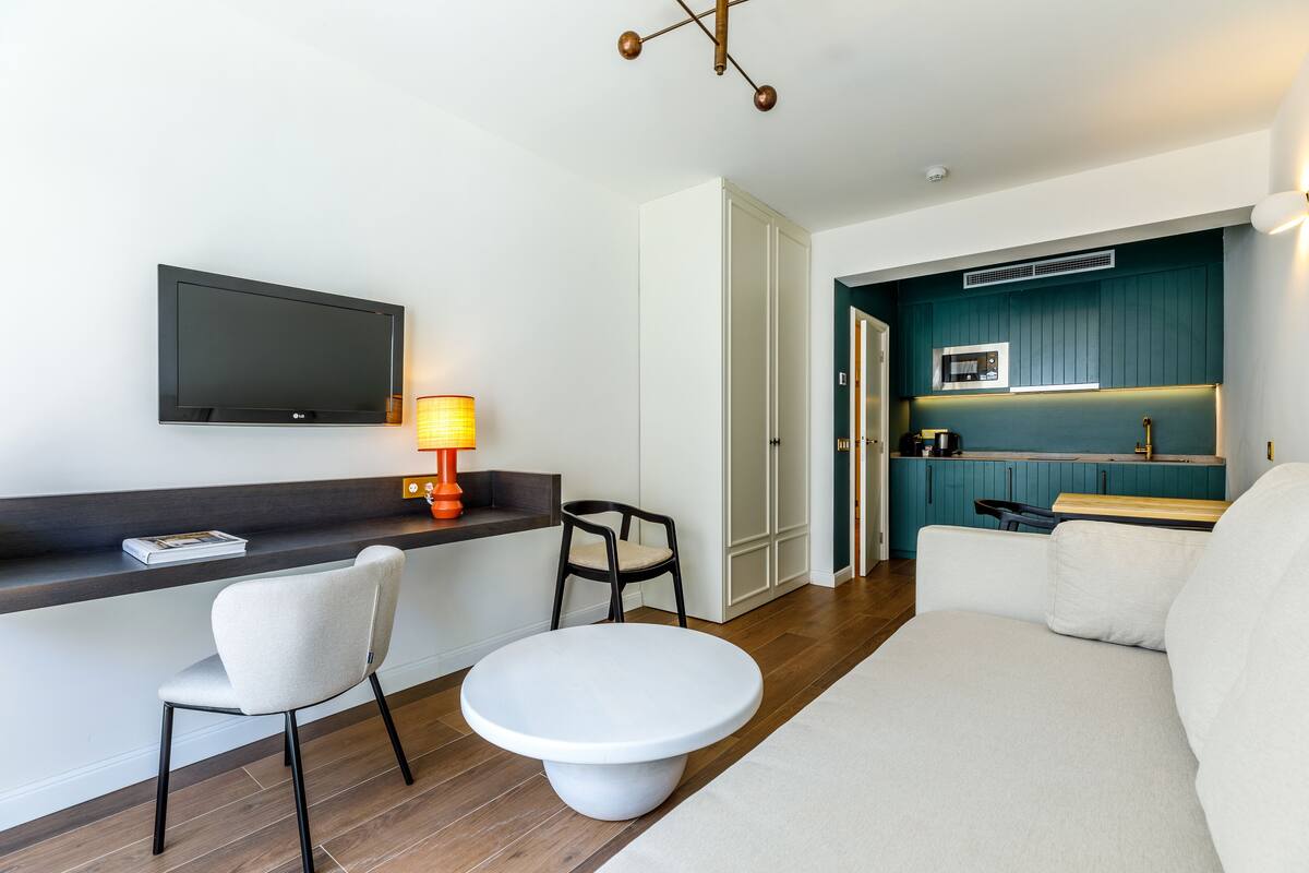 A modern living room with a flat-screen TV, a round white table, two chairs, a sofa, and a kitchen area with teal cabinetry.