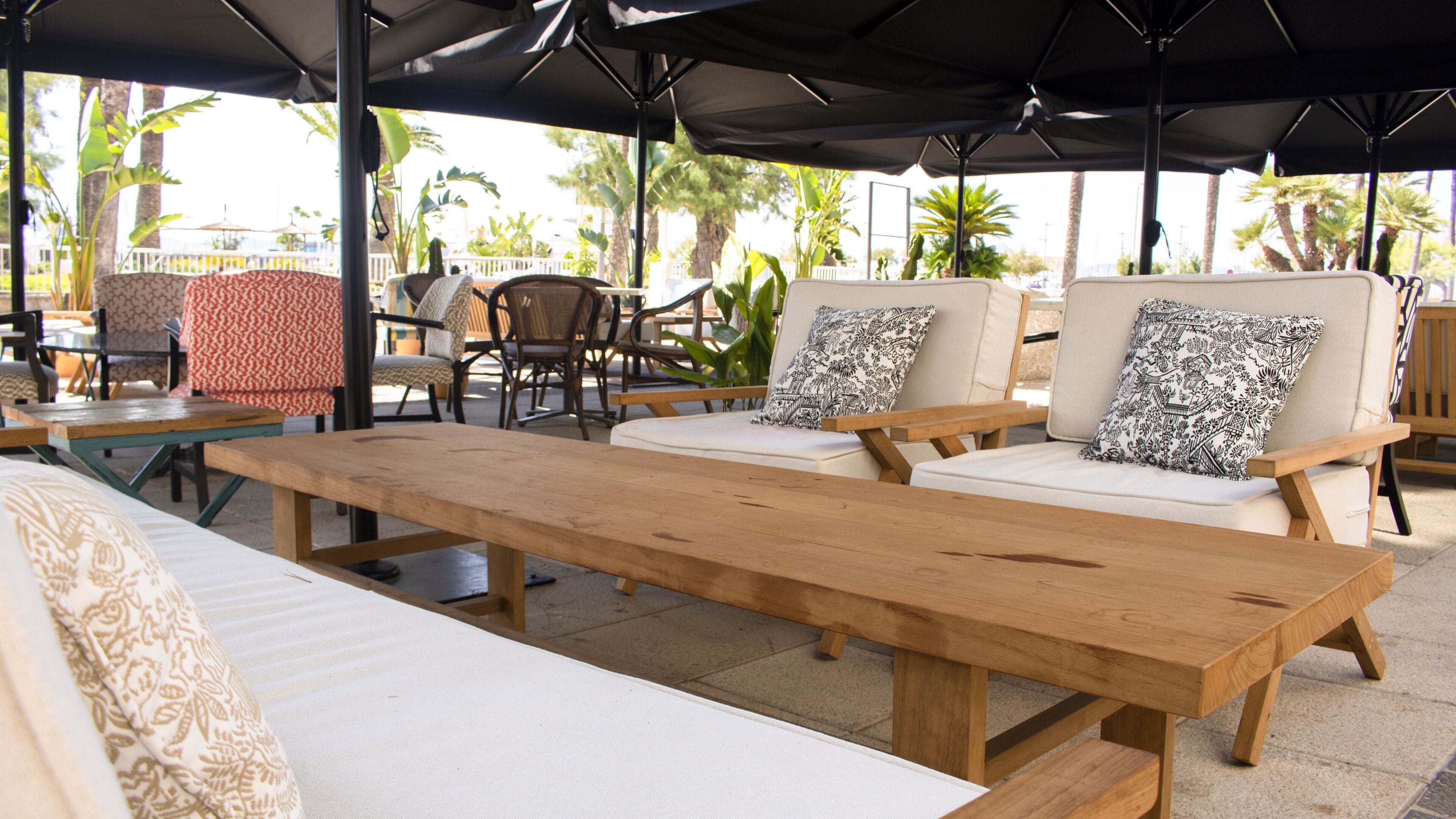 A wooden bench with two cushioned chairs, a patterned pillow, and a table under a shaded area.