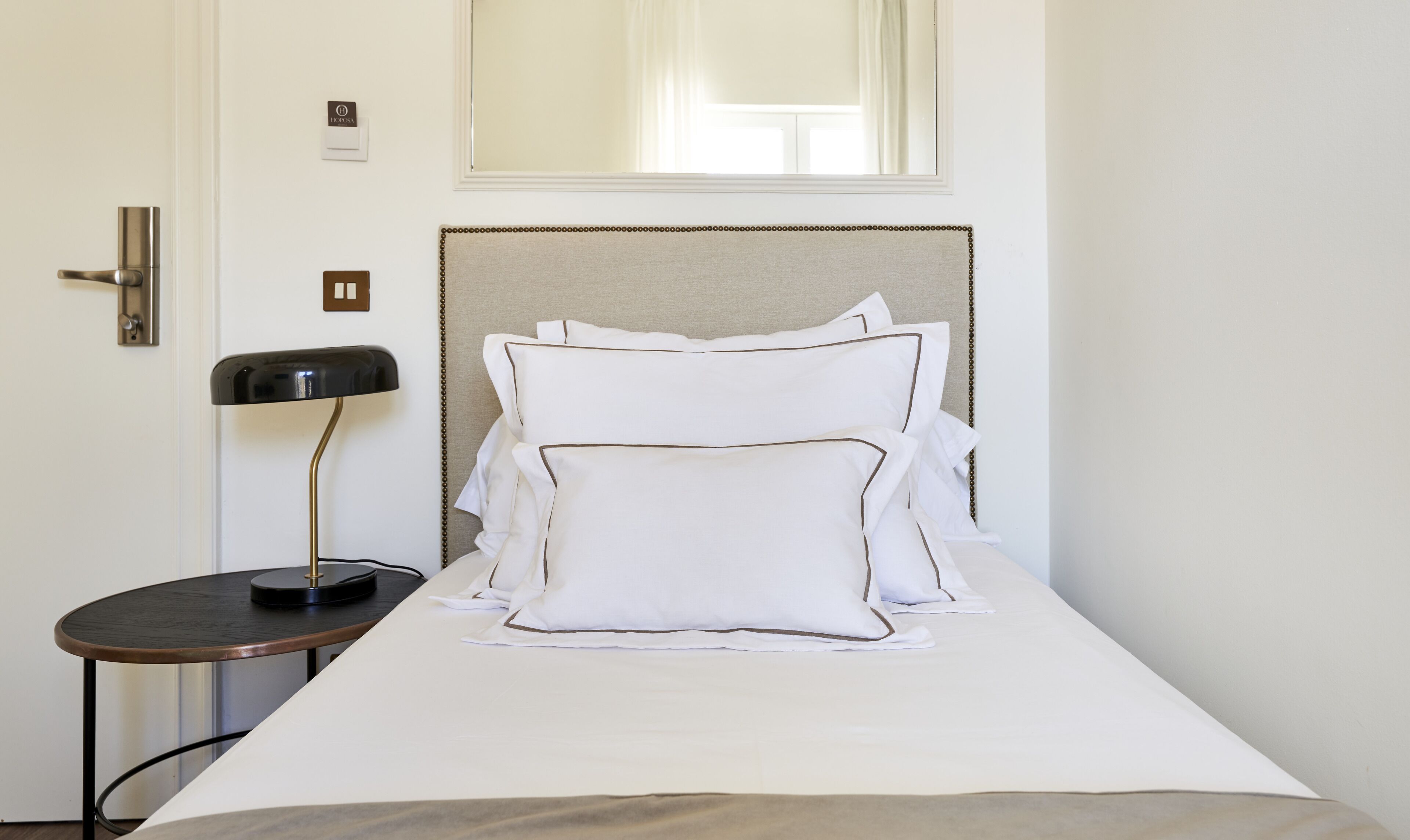 A neatly made bed with white linens and pillows, a bedside table with a lamp, and a door with a handle.