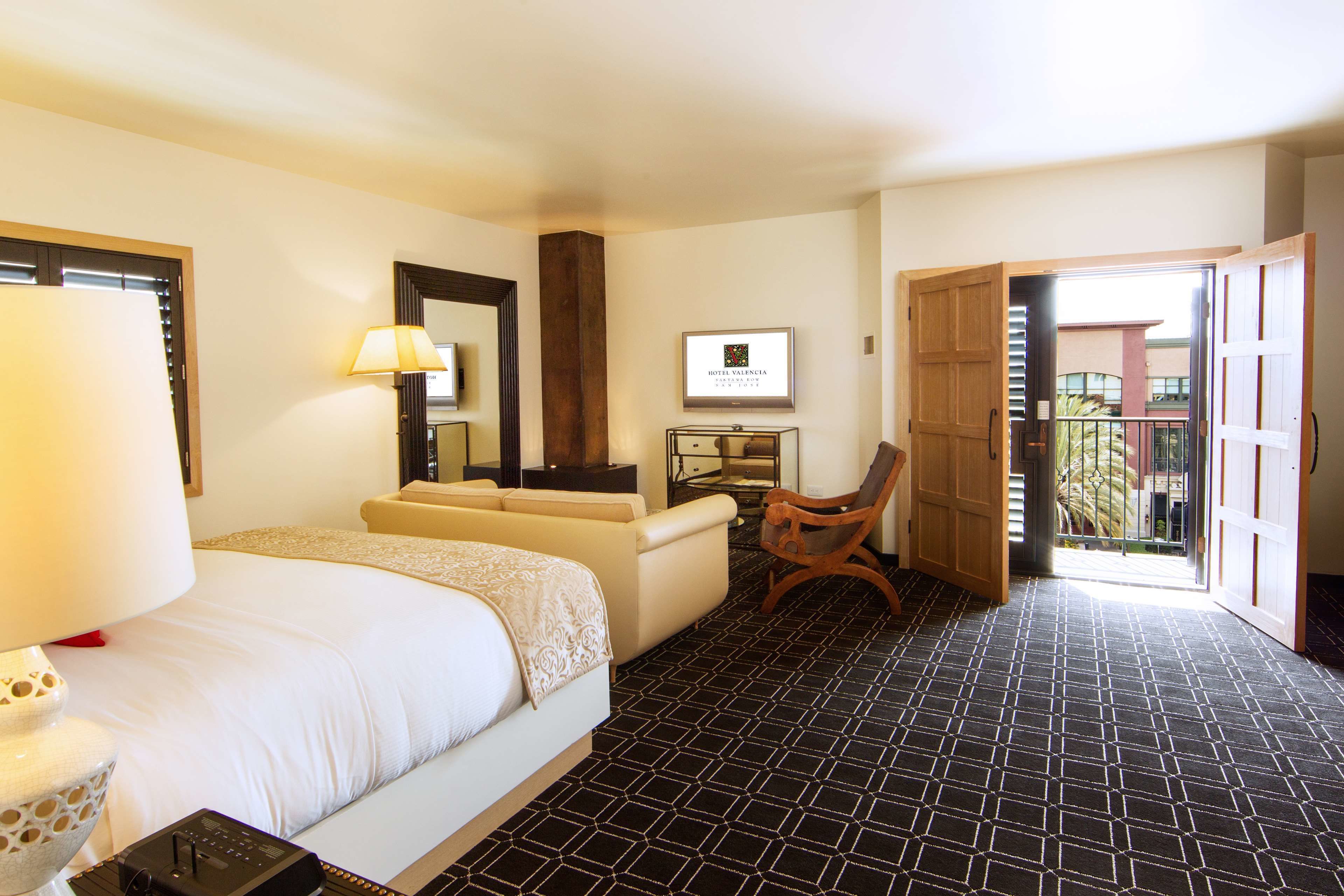 A hotel room with a bed, a desk, a chair, a lamp, a mirror, and a framed sign on the wall.