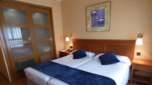 A double bed with a blue bedspread, wooden headboard, two bedside tables with lamps, a wooden wardrobe with frosted glass doors, and a framed artwork on the wall. - Eco Via Lusitana (Madrid)