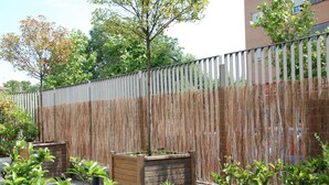 A wooden fence with vertical slats and woven panels, flanked by planter boxes with green plants. - Eco Via Lusitana (Madrid)