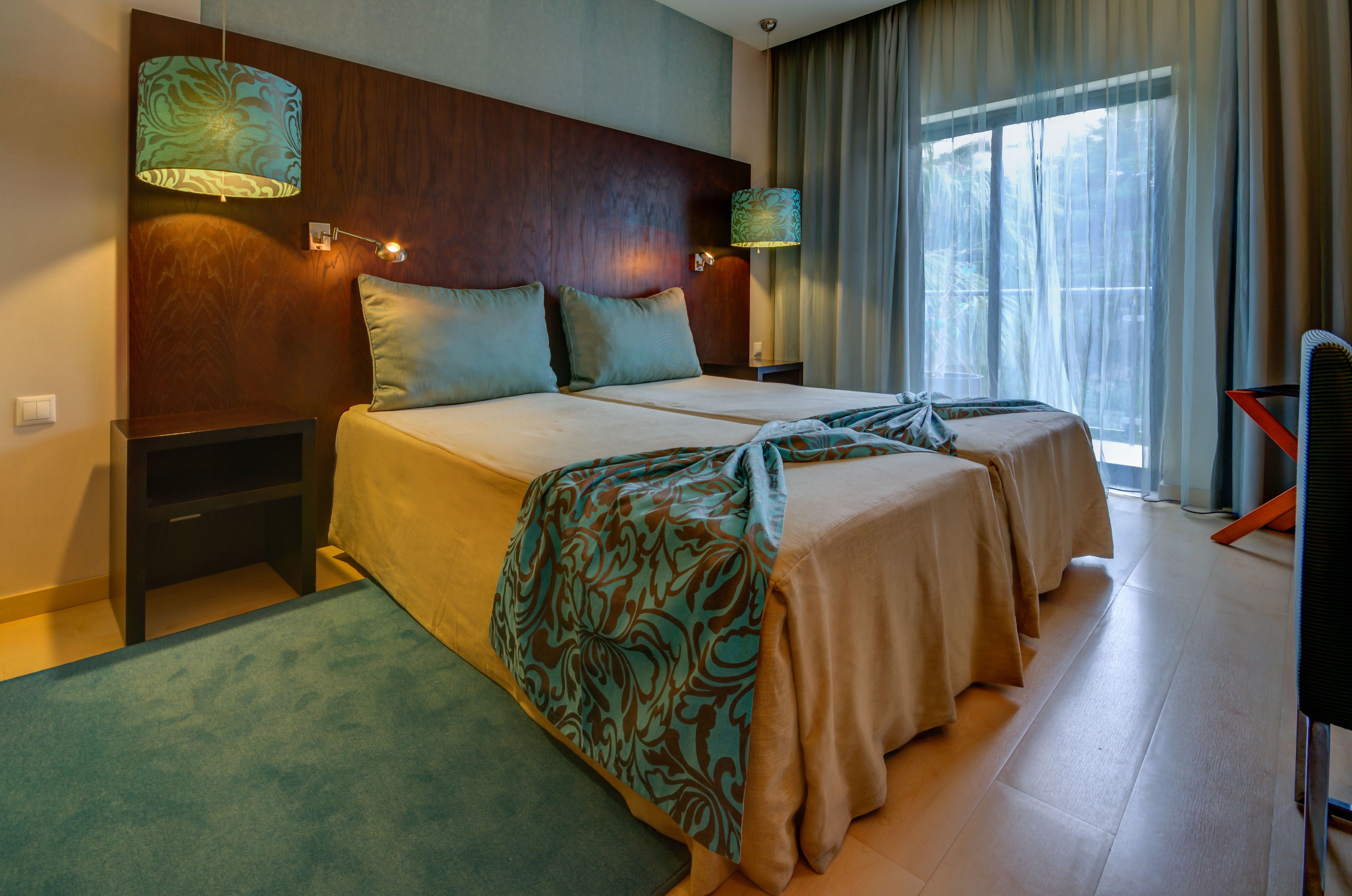 A hotel room with two beds, a wooden headboard, a large window with curtains, and a blue carpet.
