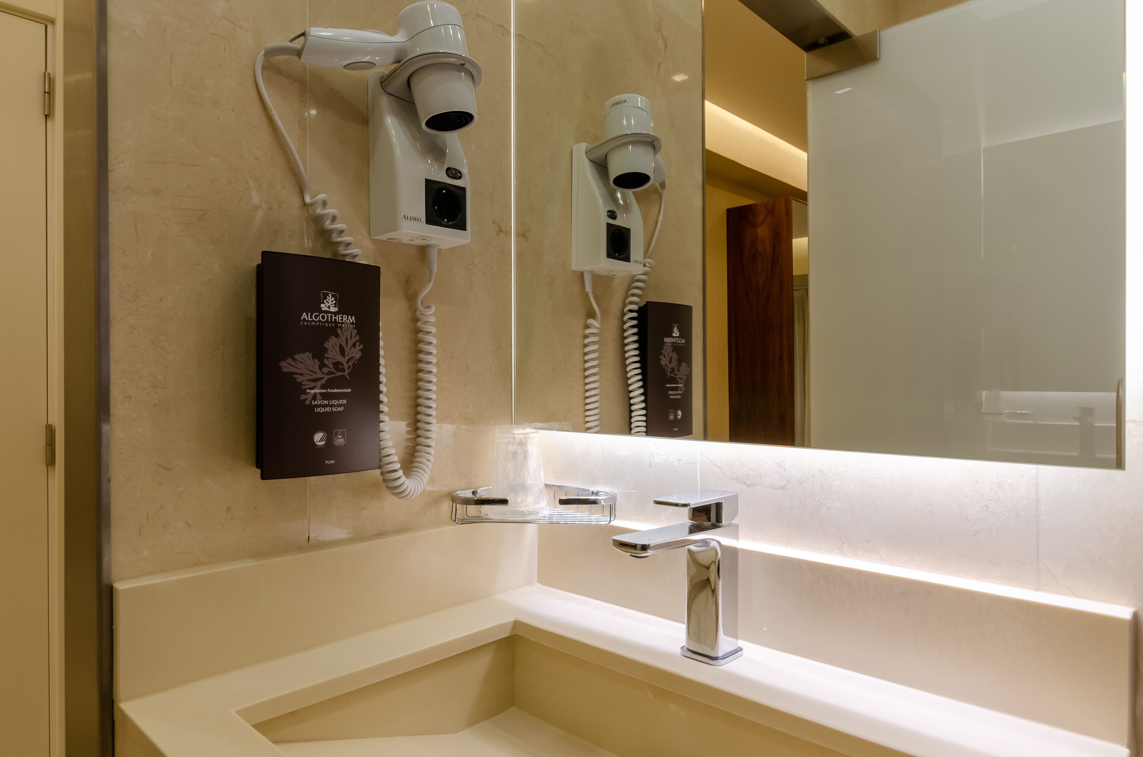 A modern bathroom with a large mirror, a built-in hair dryer, and a bottle of Algotherm liquid soap.