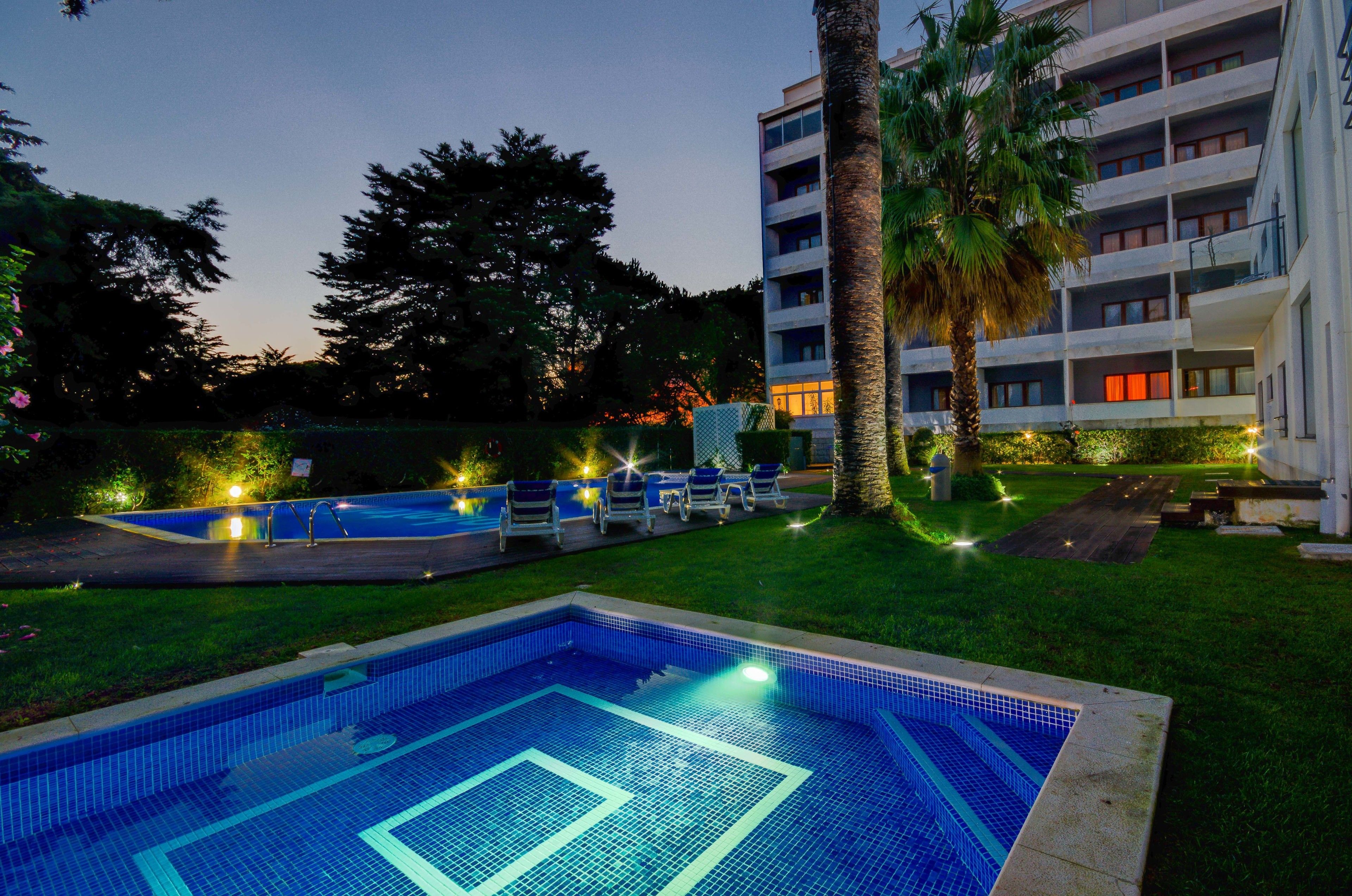 A swimming pool with lounge chairs and a palm tree, surrounded by a building with multiple floors and balconies.