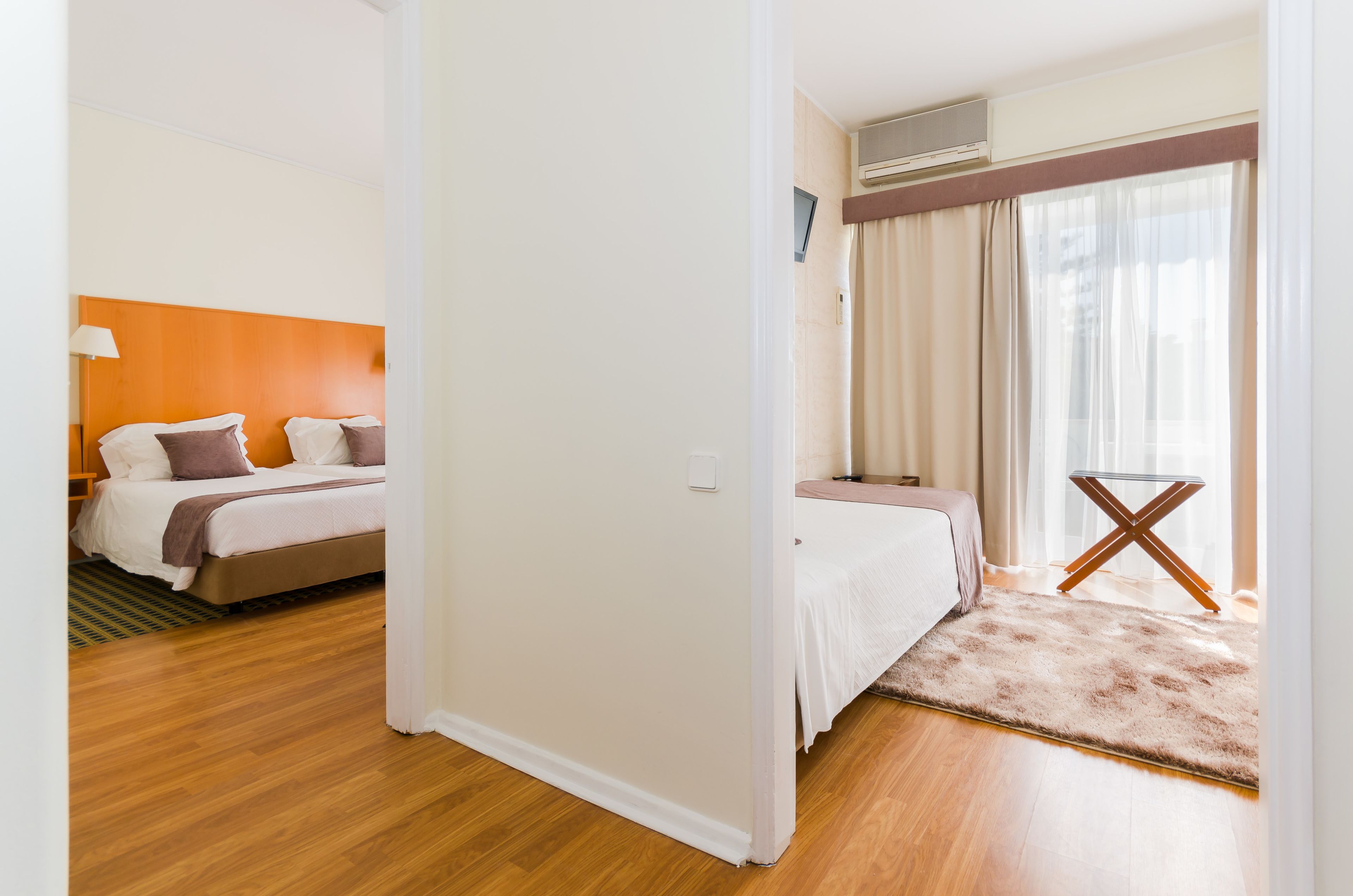 A hotel room with a wooden floor, a bed with white and brown bedding, a small table with a cross-back design, and a window with sheer curtains.
