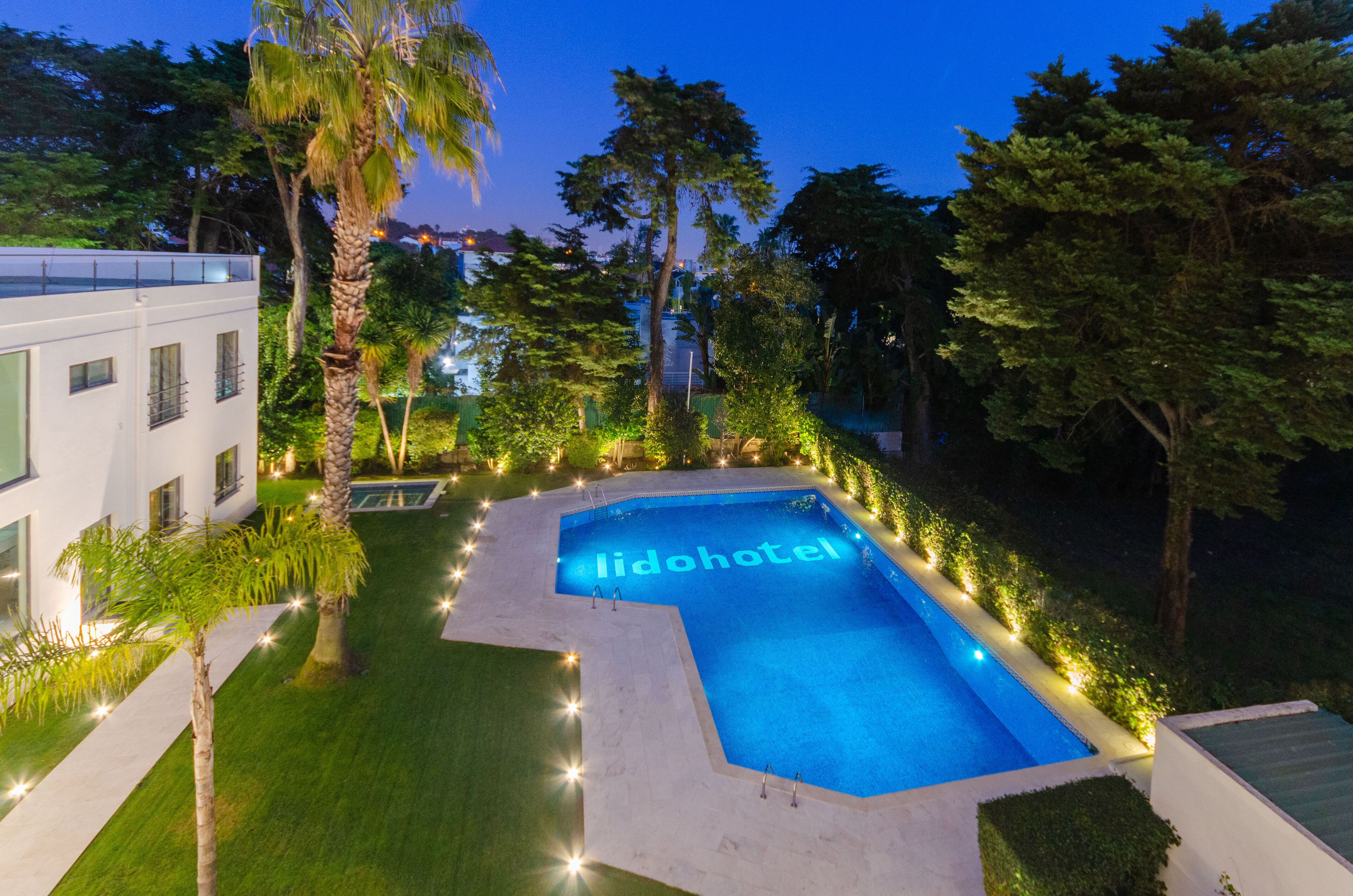 Una piscina con la inscripción 'lodohotel', rodeada de follaje bien cuidado y un edificio moderno.