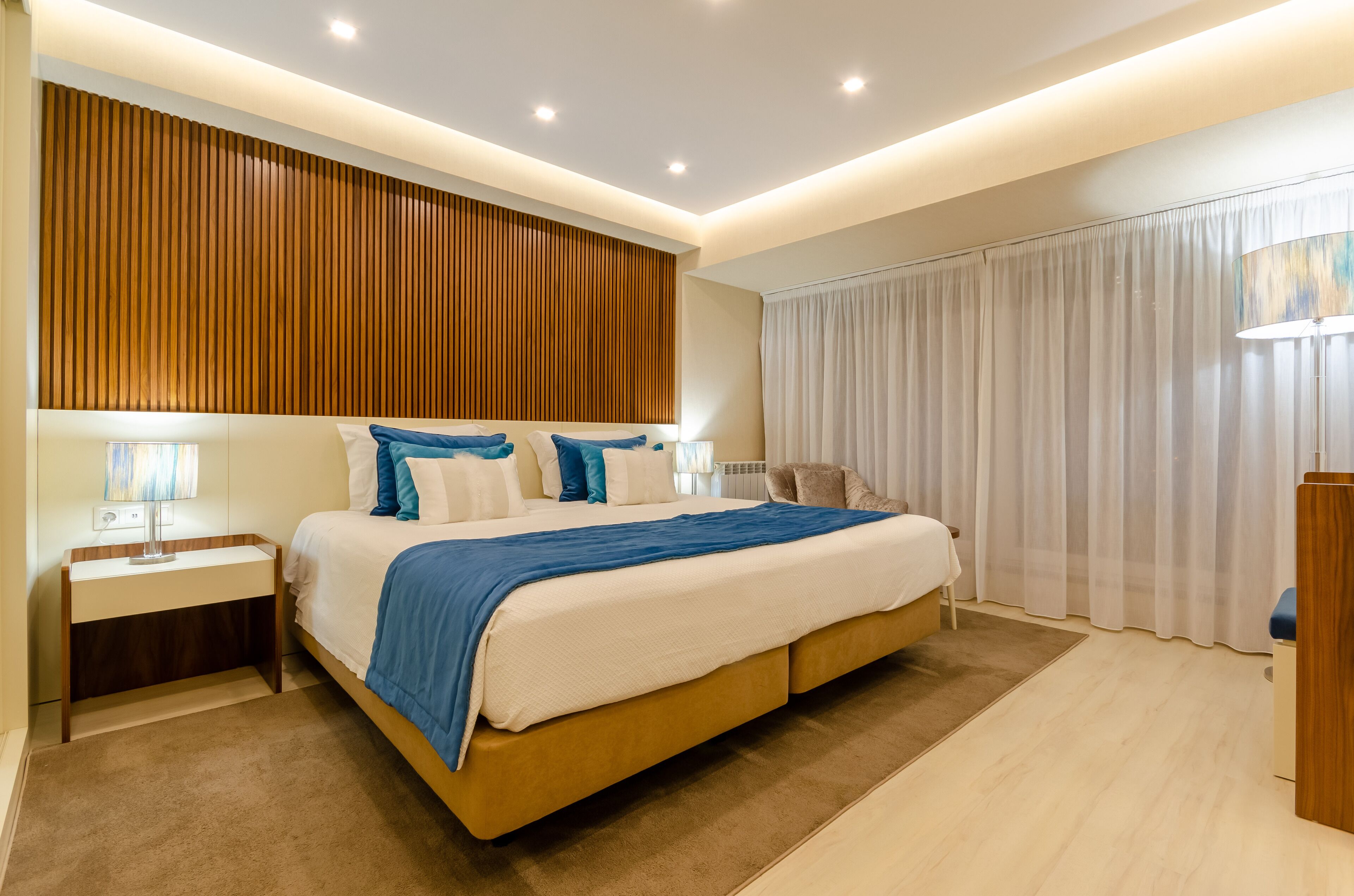 A modern bedroom with a large bed, bedside tables, a wooden accent wall, and a large window with curtains.