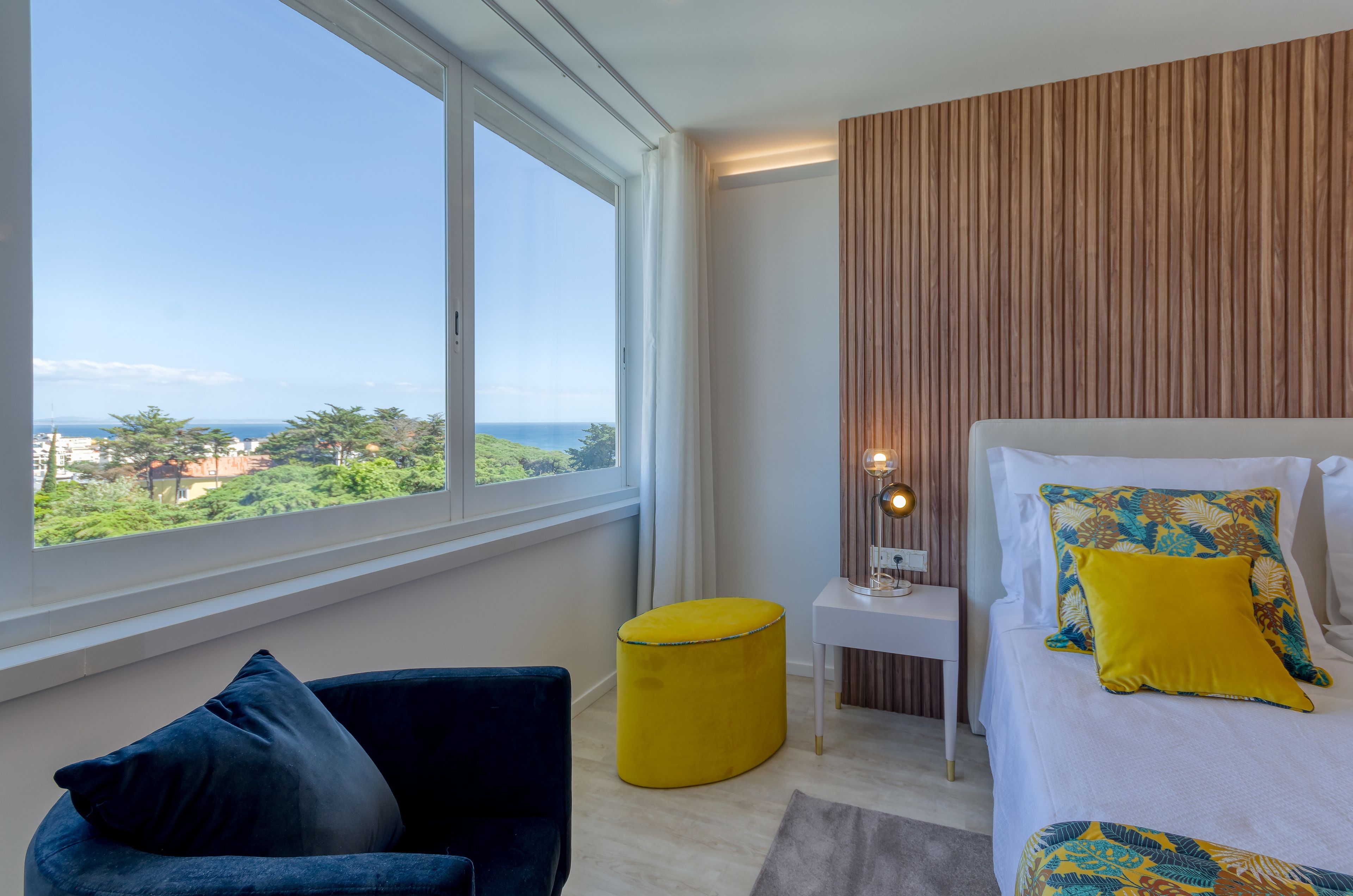 Ein modernes Schlafzimmer mit großem Fenster, einem Bett mit gemusterten Kissen, einem kleinen Beistelltisch und einem gelben Hocker.