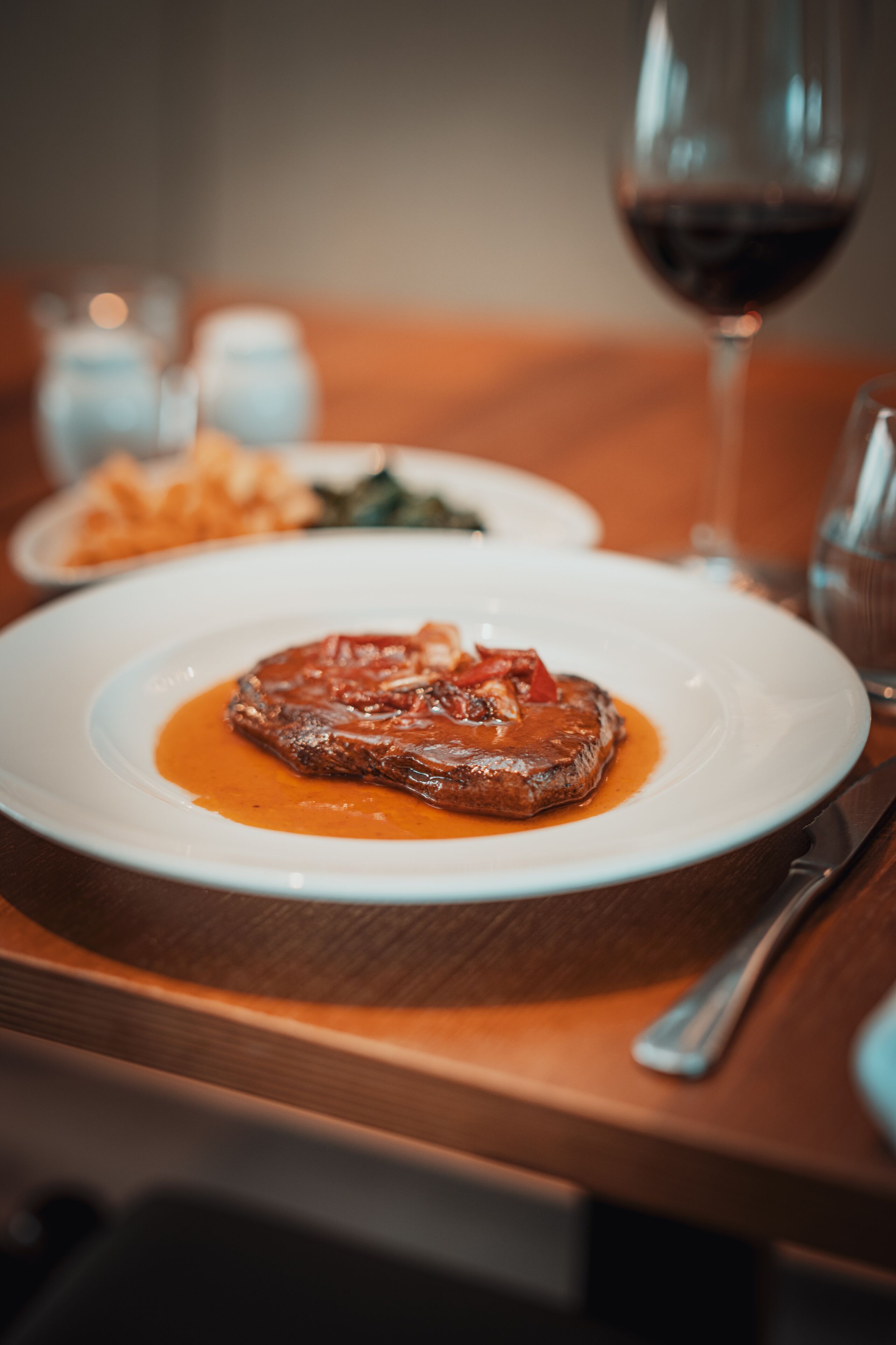 A steak with sauce on a white plate, a glass of red wine, and a fork on a wooden table.