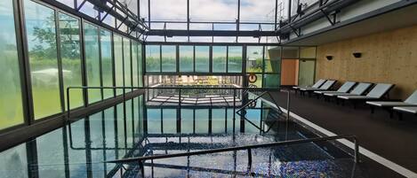 An indoor swimming pool with glass walls, a wooden deck, and lounge chairs.
