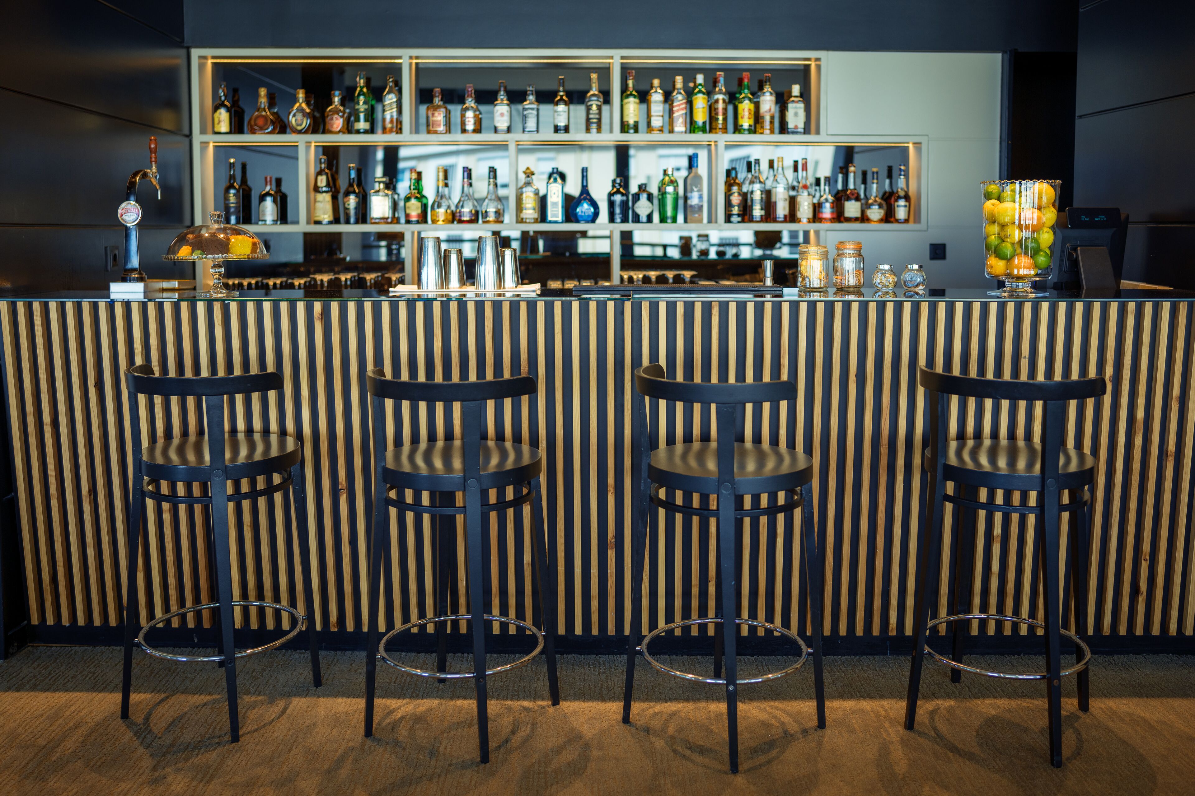 Un espace bar avec un comptoir en bois à rayures, des tabourets en métal et des étagères remplies de bouteilles d’alcool.