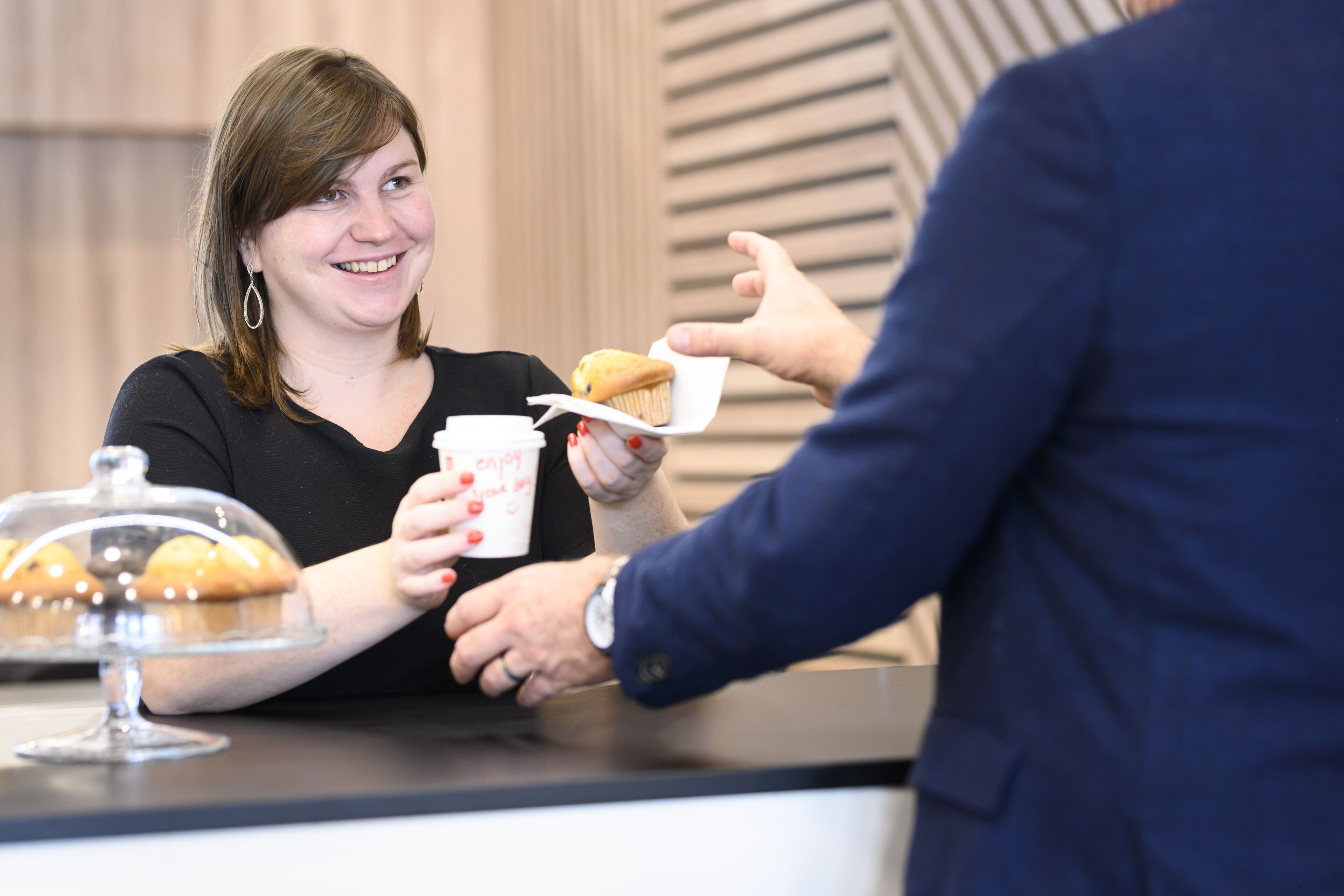 Una persona sosteniendo un muffin y una taza con el texto 'enjoy your day' sobre un mostrador con una variedad de muffins.