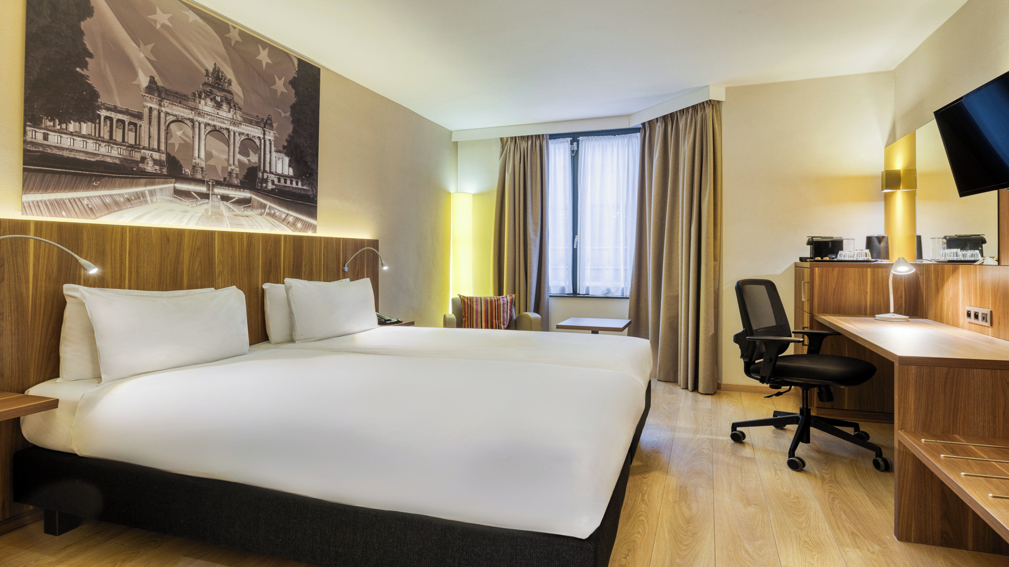 A hotel room with a large bed, a desk, a chair, a TV, and a window with curtains.