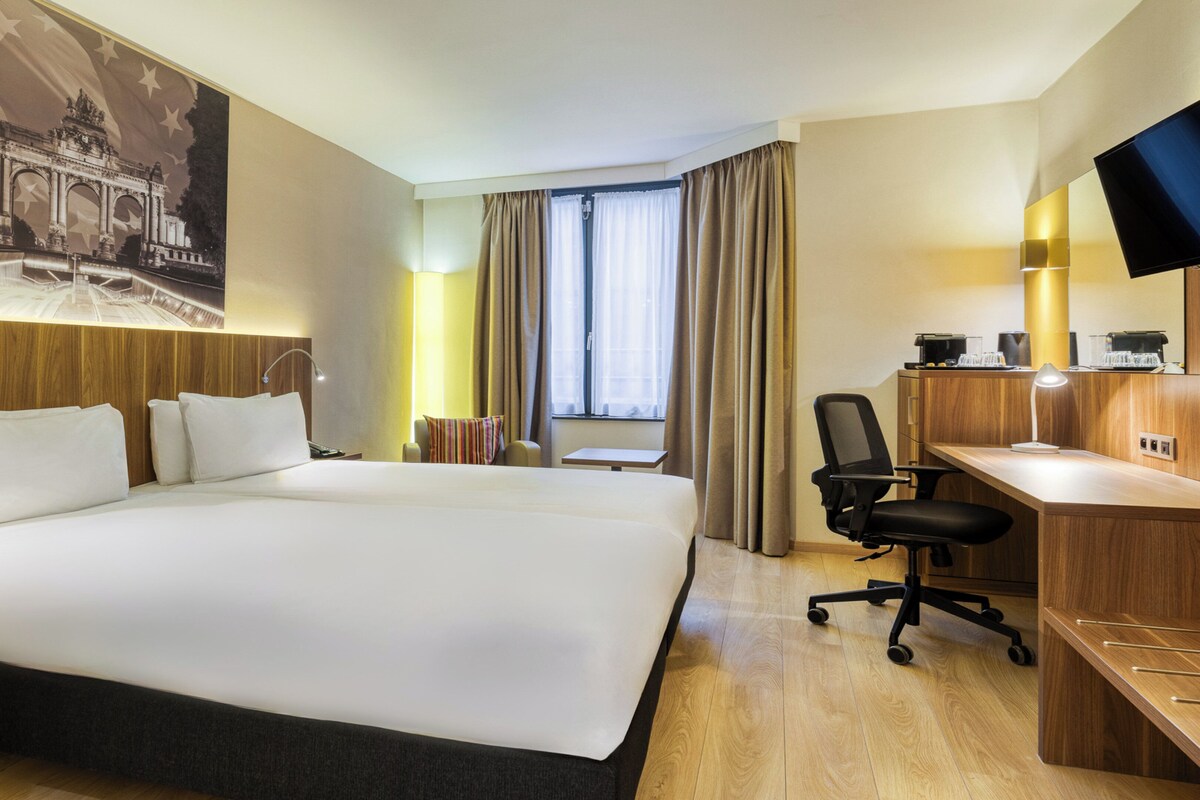 A hotel room with a large bed, a desk, a chair, a TV, and a window with curtains.