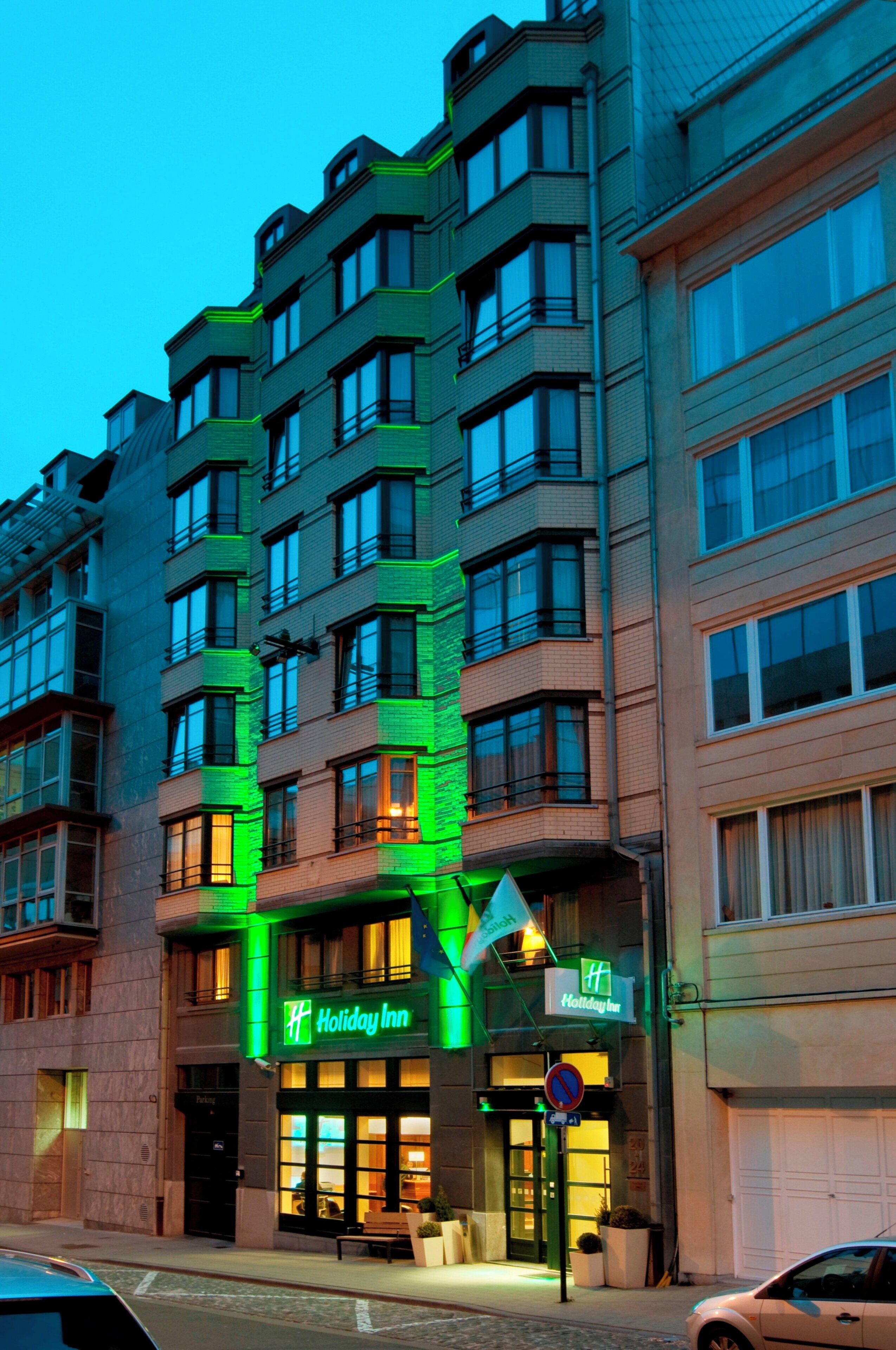 Un immeuble à plusieurs étages, sur lequel on peut voir l’enseigne d’un hôtel Holiday Inn, se distinguant par son architecture moderne, ses grandes fenêtres et sa façade illuminée de lumière verte.
