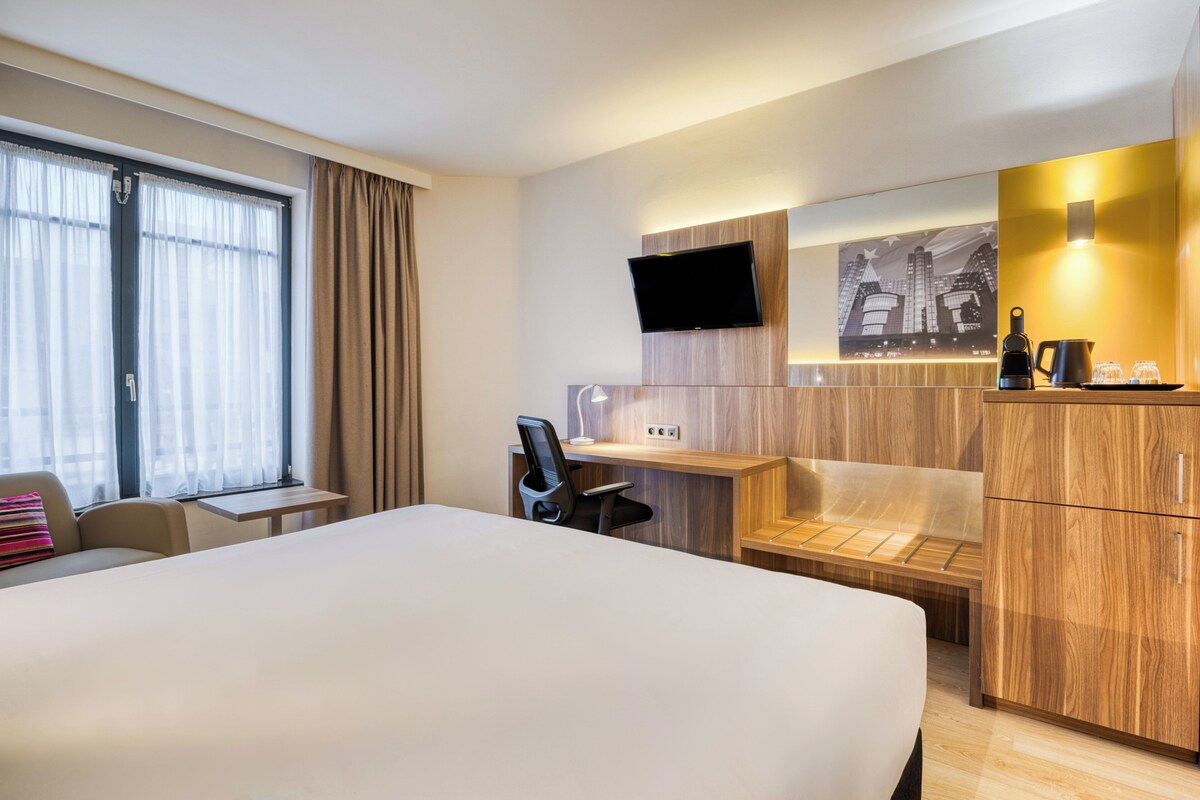 A hotel room with a large bed, a desk with a chair, a TV mounted on the wall, and a wooden cabinet.