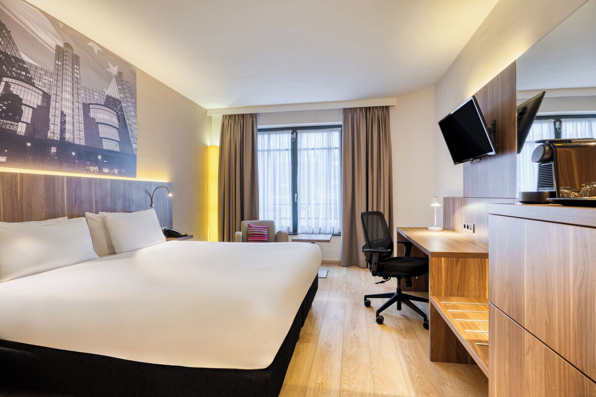 A hotel room with a large bed, a desk with a computer, a chair, a TV mounted on the wall, and a window with curtains.