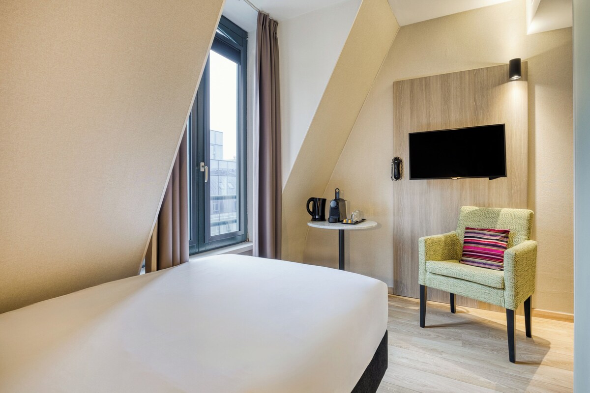 A modern hotel room with a large bed, a small round table with a coffee maker, a chair with a colorful cushion, and a flat-screen TV mounted on the wall.