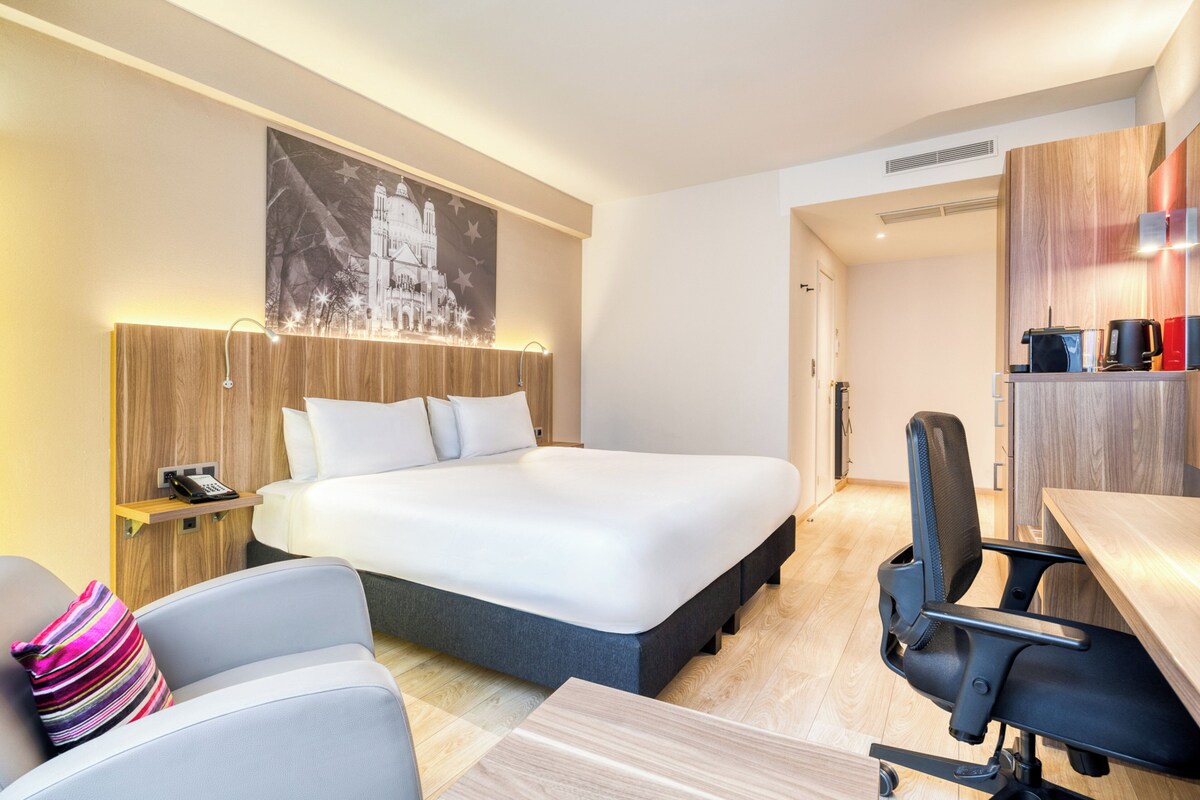A modern hotel room with a large bed, a desk, a chair, and a small table.