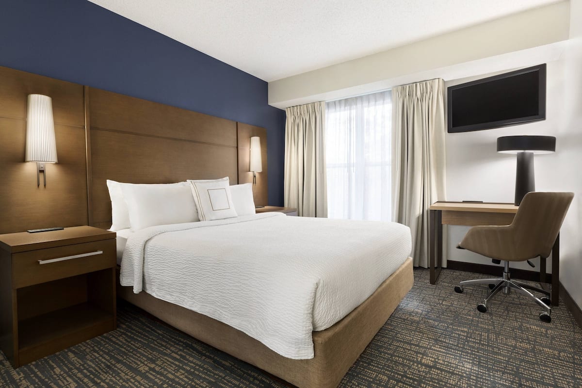 A hotel room with a large bed, a desk with a chair, a flat-screen TV, and a nightstand with a lamp.