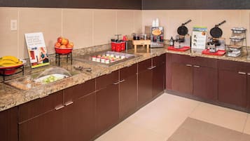 A kitchen area with a granite countertop, a fruit bowl, a fruit tray, a waffle maker, and a coffee station.
