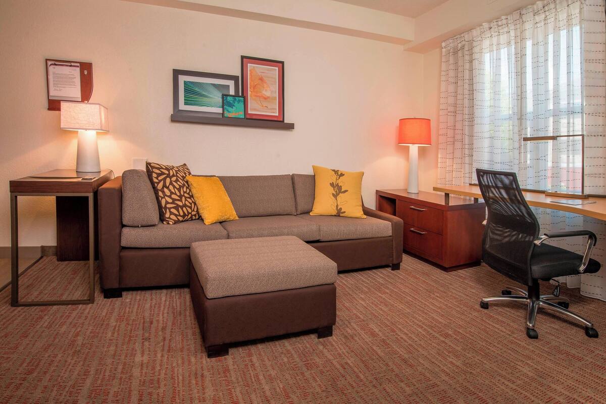 Quarto de hotel com sofá, apoio para os pés, escrivaninha, cadeira e janela com cortinas.