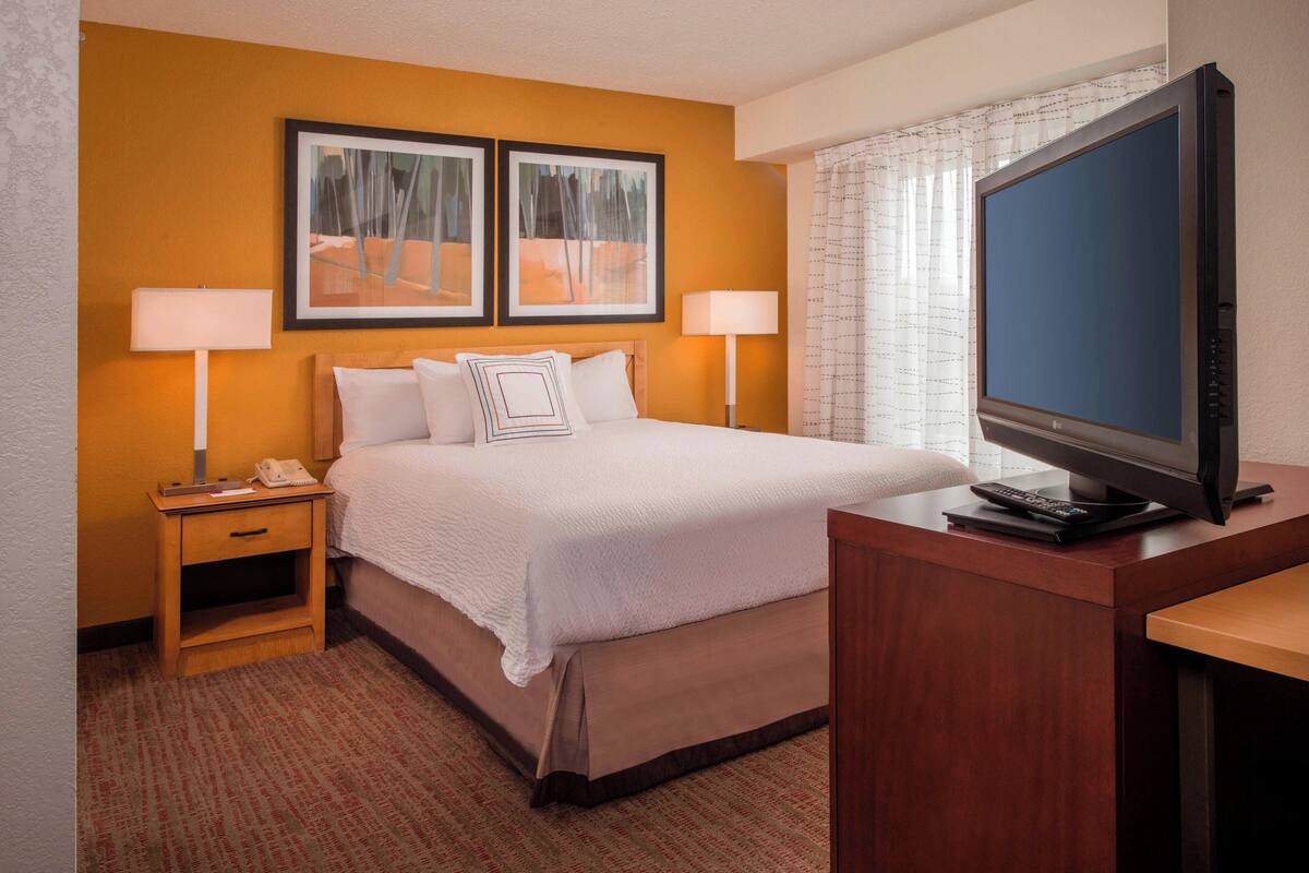 Quarto de hotel com cama, luminárias de cabeceira, televisão e obras de arte na parede.