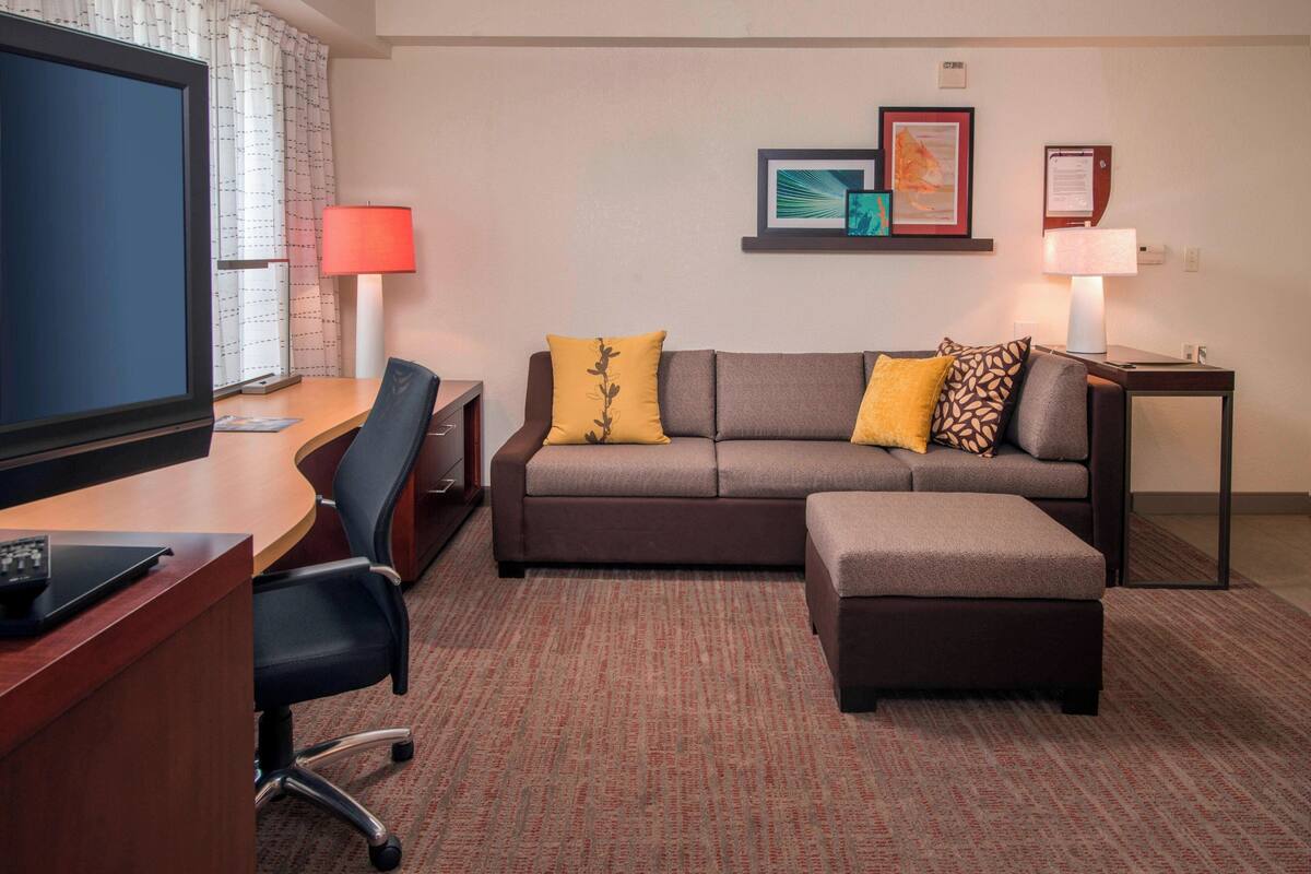 Quarto de hotel com uma televisão de tela grande, uma escrivaninha com um laptop, um sofá com dois almofadas amarelas, uma mesinha de cabeceira com uma luminária e um quadro na parede.