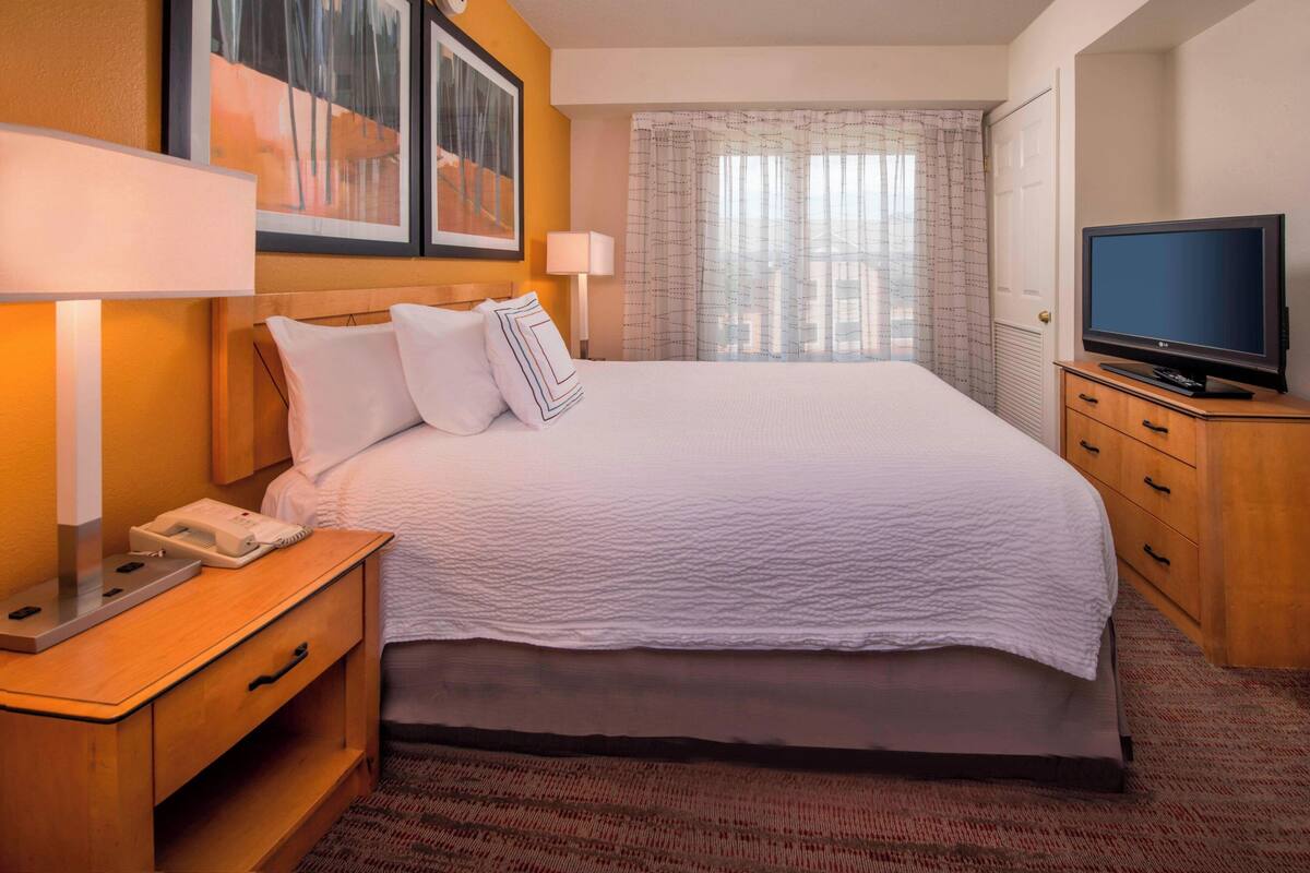 Quarto de hotel com uma cama grande, uma televisão em uma cômoda de madeira, abajur, telefone e uma obra de arte na parede.