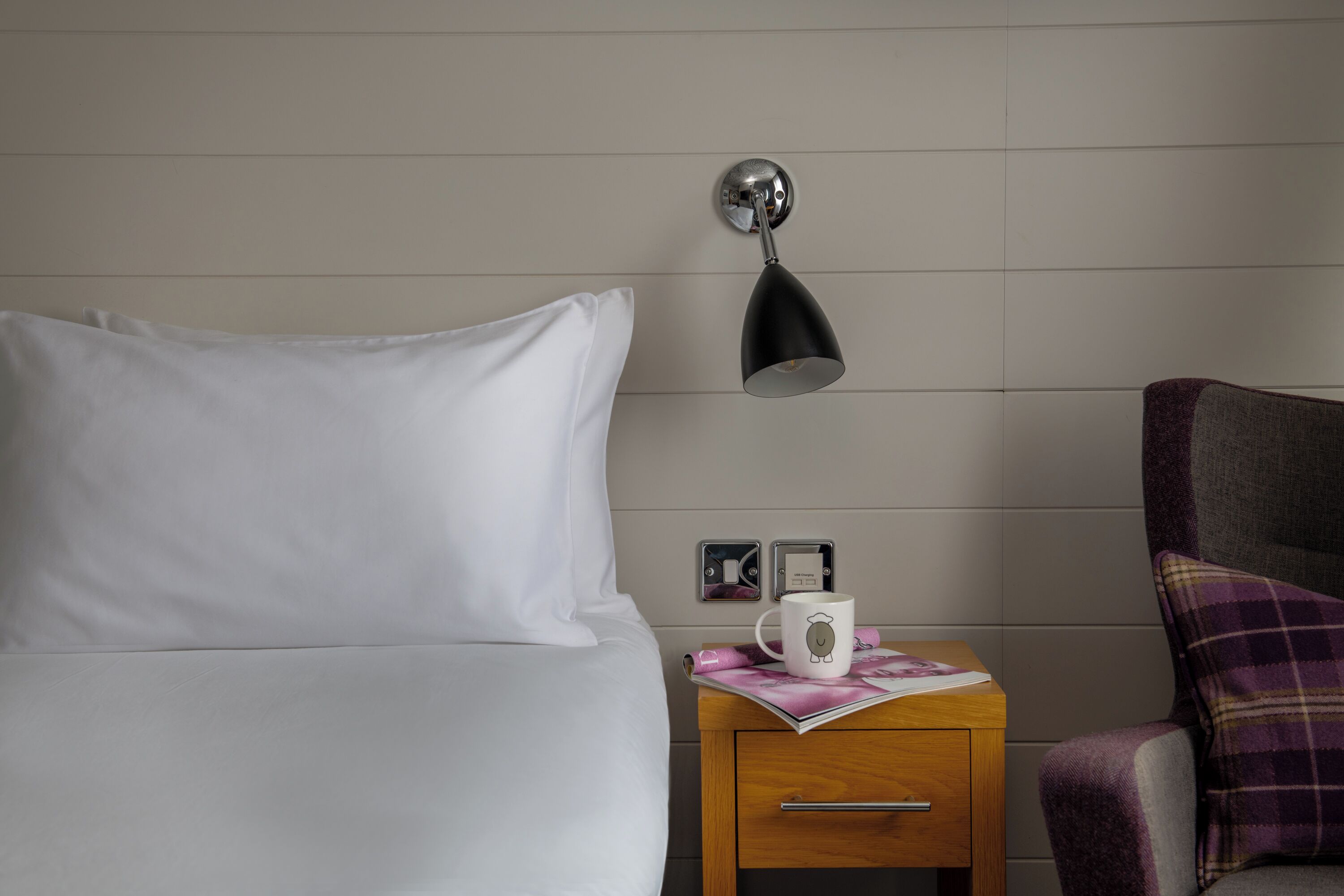 A neatly made bed with white sheets, a bedside table with a lamp, a mug, and a magazine.