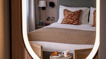 A hotel room with a bed, a bedside table with a lamp, a mirror, and a small table with a cup and a notebook.