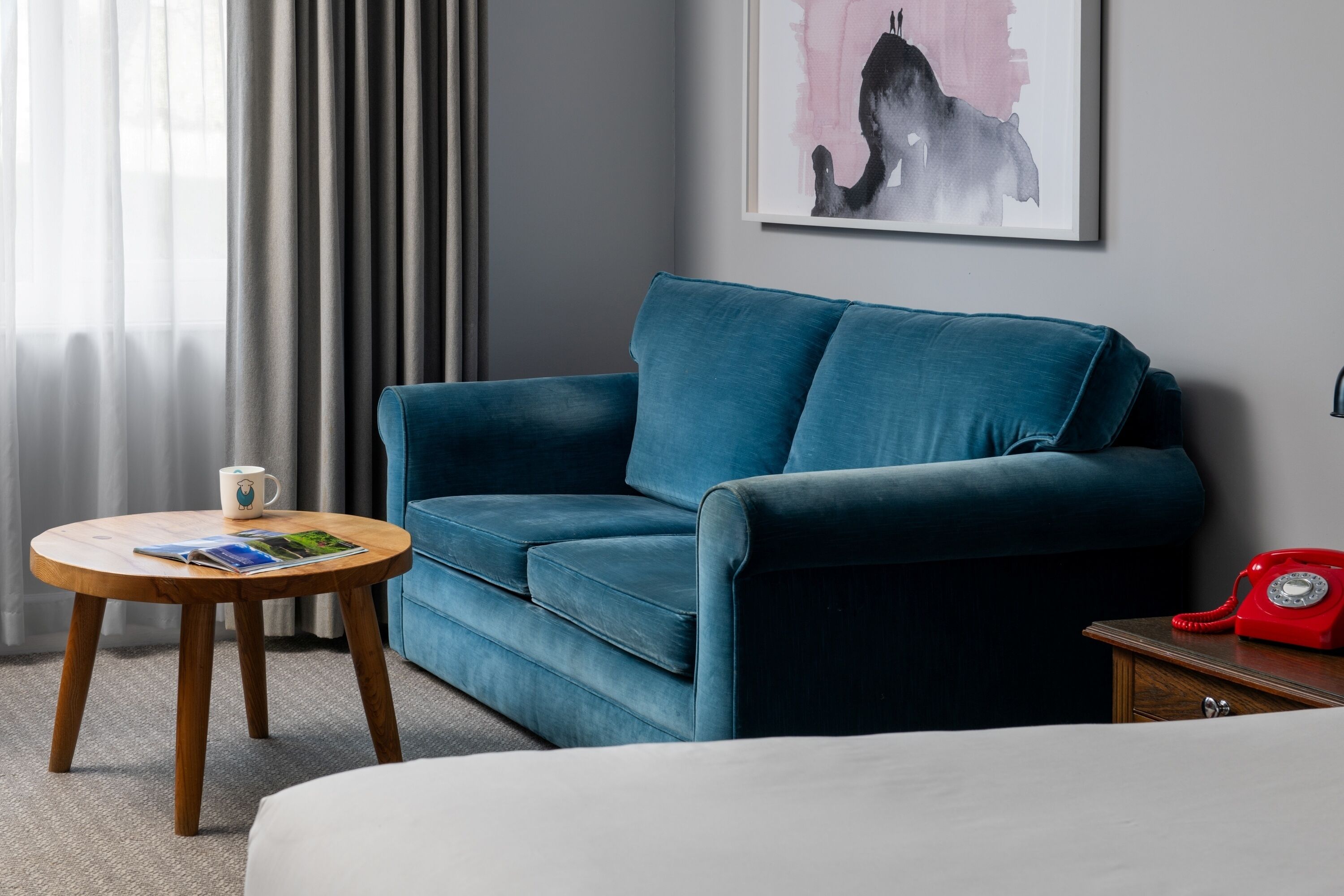 A modern hotel room with a blue sofa, a wooden side table with a magazine, a red rotary phone, and a framed artwork on the wall.