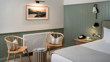 Une chambre d’hôtel avec un lit, deux chaises en bois, une petite table avec une lampe, un tableau encadré au mur et un radiateur.