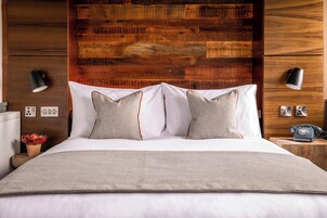 A neatly made bed with white linens and two pillows, a wooden headboard, bedside lamps, a nightstand with a telephone, and a small plant.