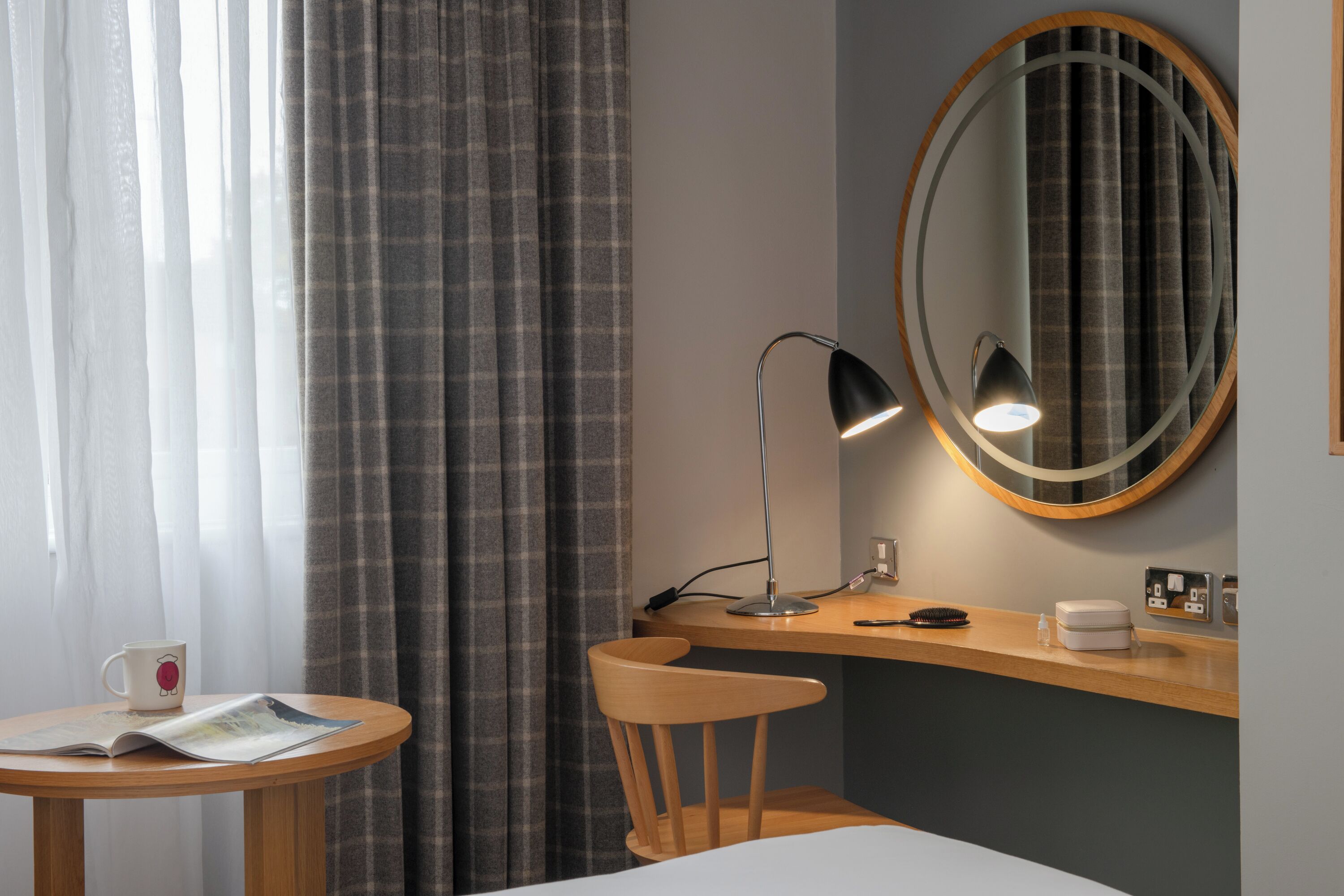 A hotel room with a wooden desk, a round mirror, a lamp, a chair, a bed, and a window with curtains.