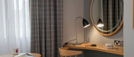 A hotel room with a wooden desk, a round mirror, a lamp, a chair, a bed, and a window with curtains.