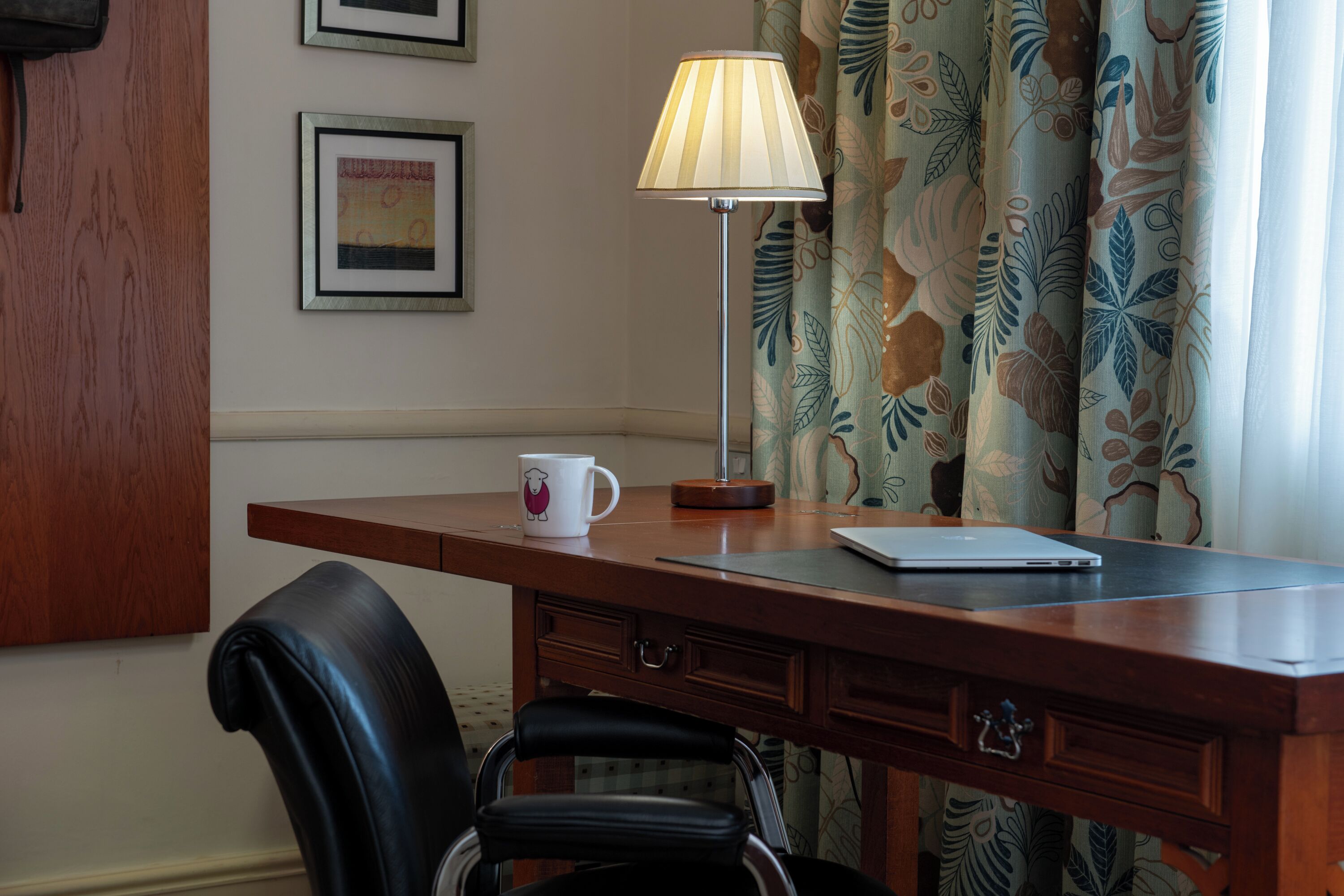 A wooden desk with a laptop, a mug, and a lamp. There is a chair and a curtain with a leaf pattern.