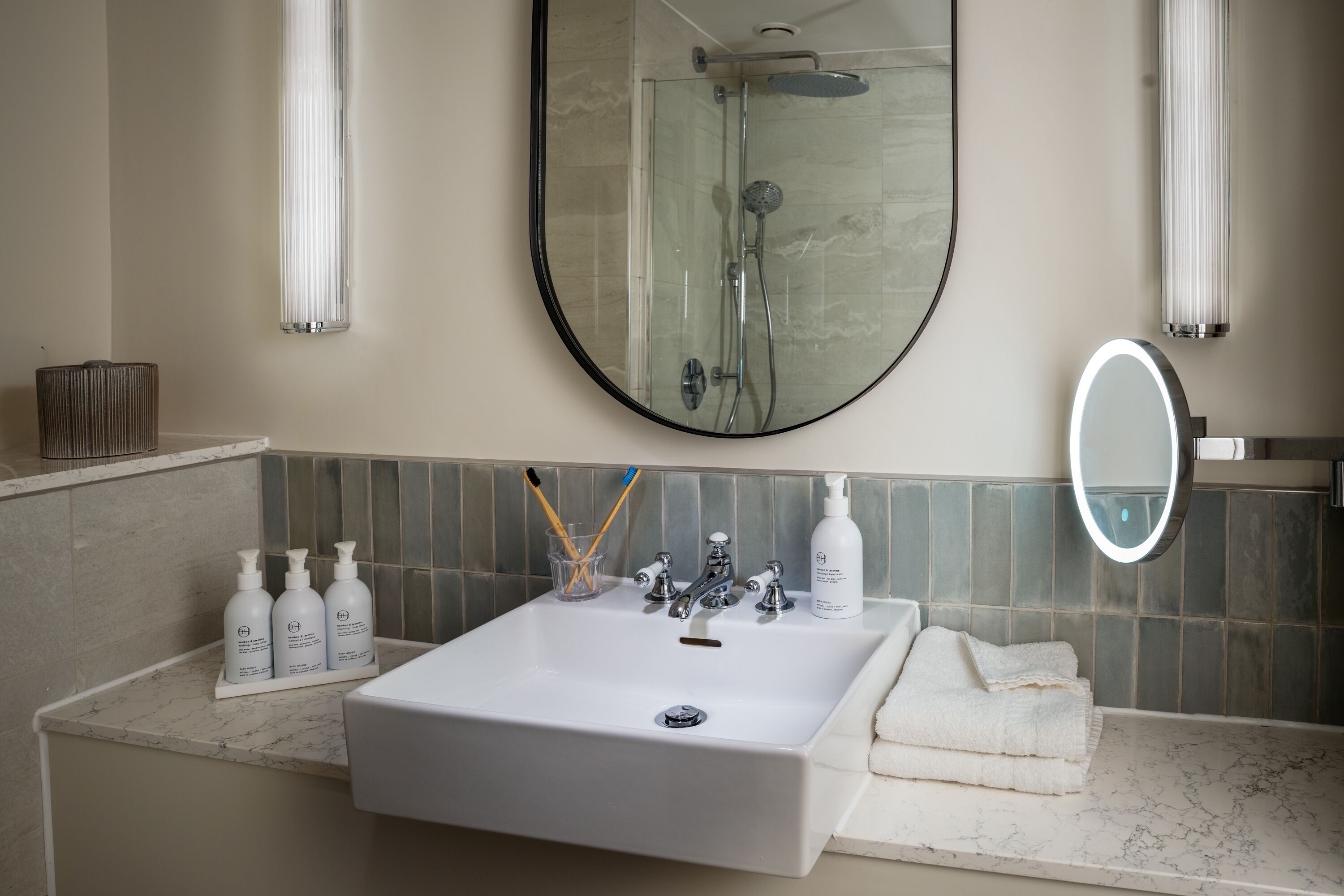 A bathroom with a white sink, a mirror, and a shower. There are toiletries on the sink and a towel on the counter.
