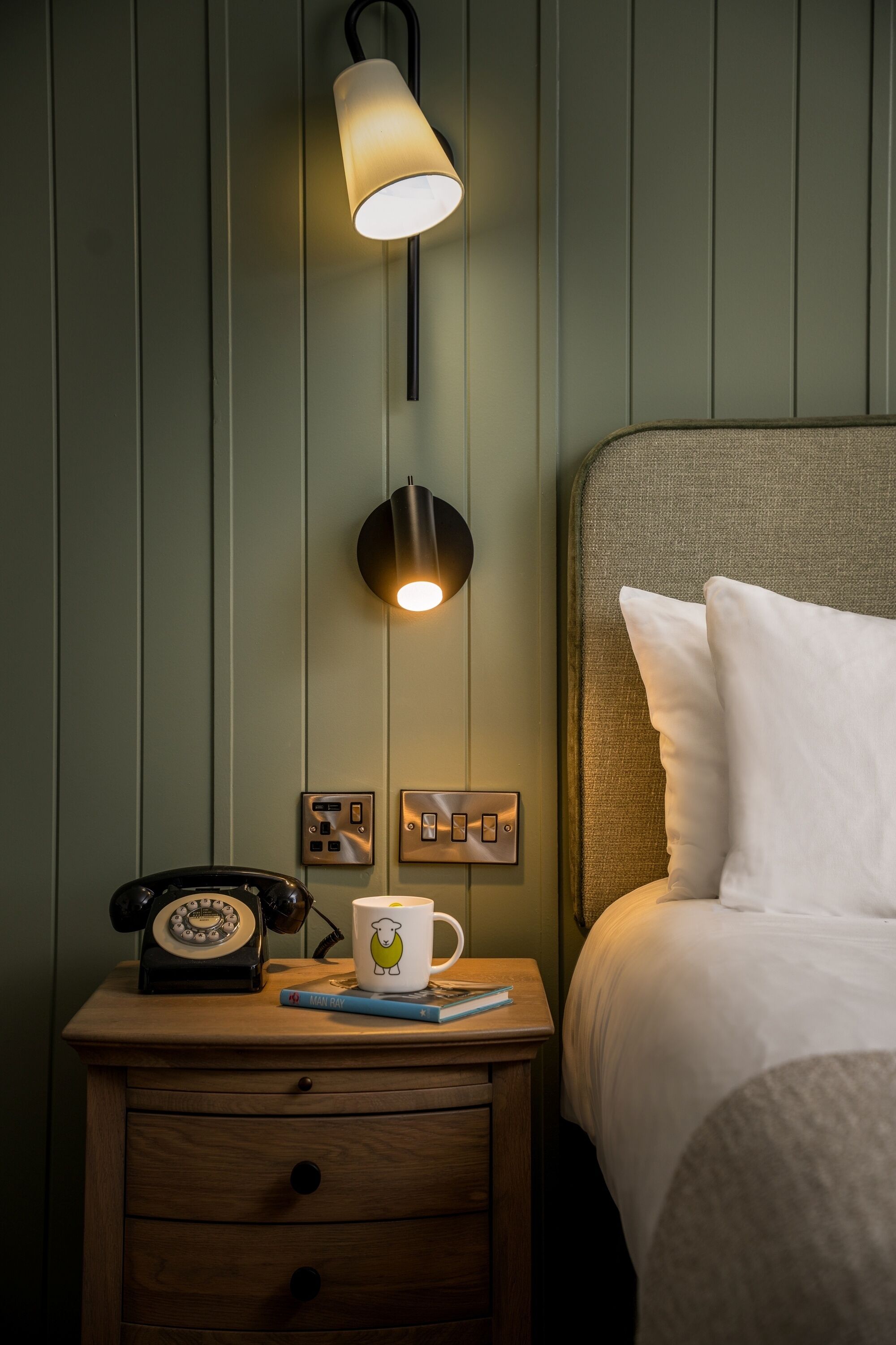 A neatly made bed with white linens, a bedside lamp, a vintage rotary phone, and a mug on a wooden nightstand.