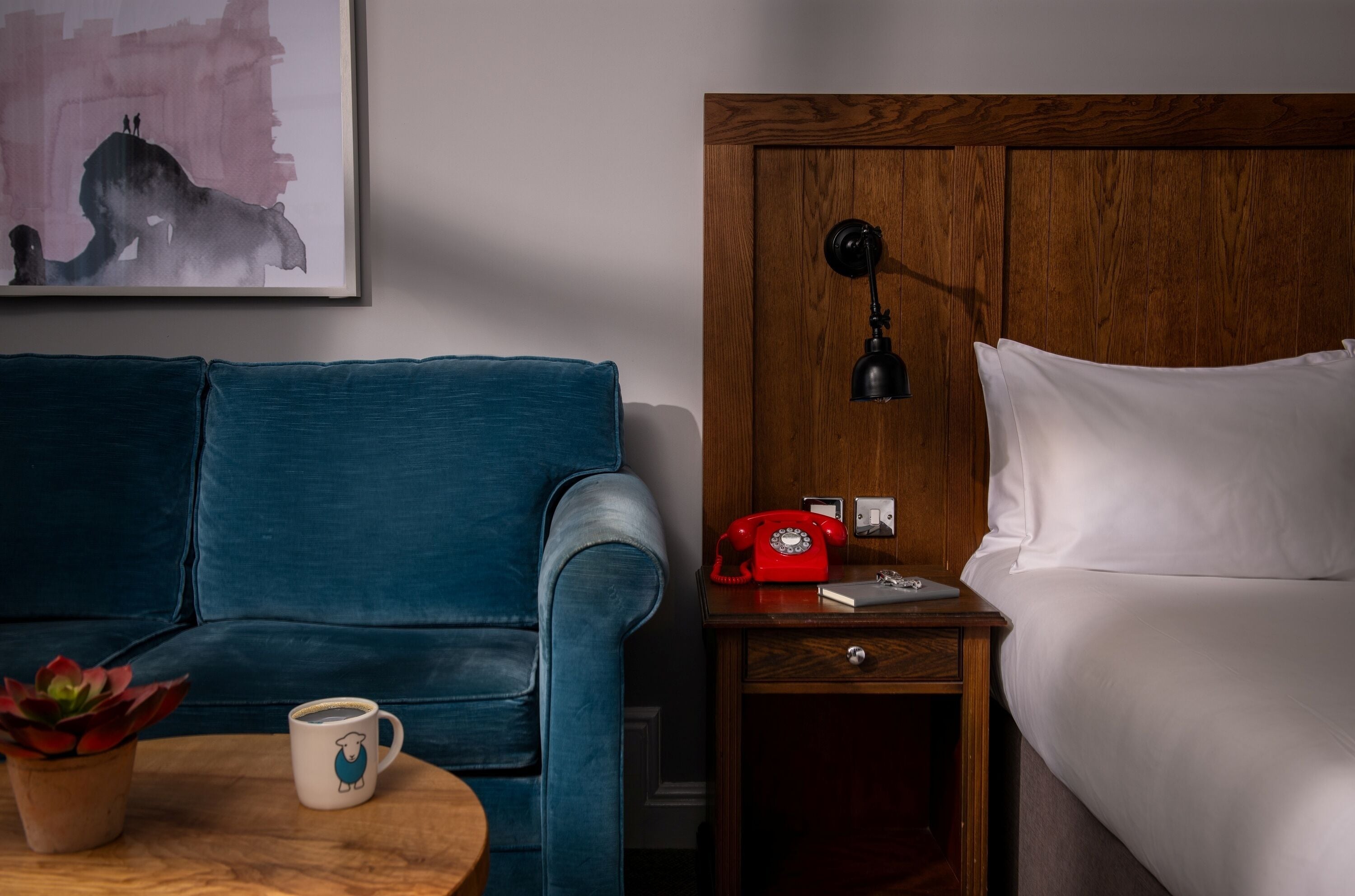 A hotel room with a blue sofa, a wooden nightstand with a red rotary phone, a bedside table with a white mug, and a painting on the wall.