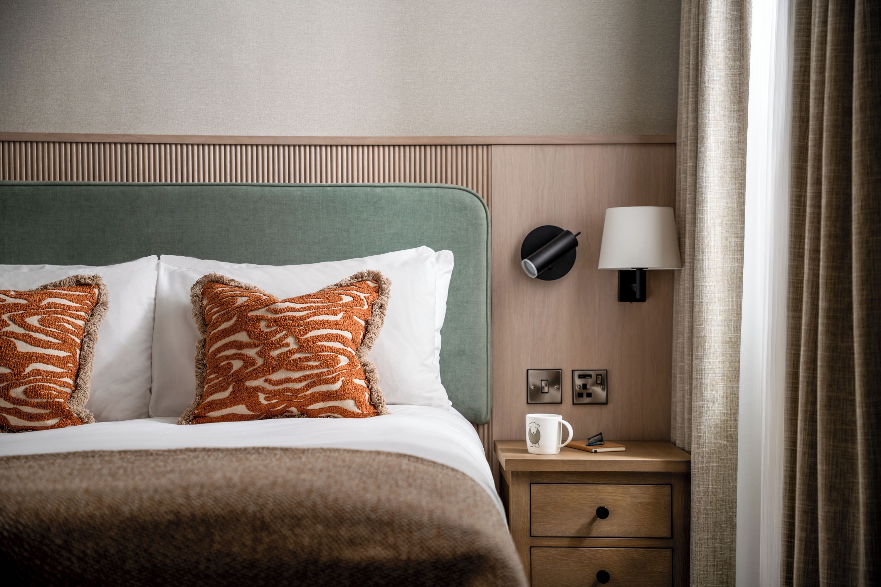 A neatly made bed with patterned pillows, a bedside table with a lamp, and a nightstand with a mug.