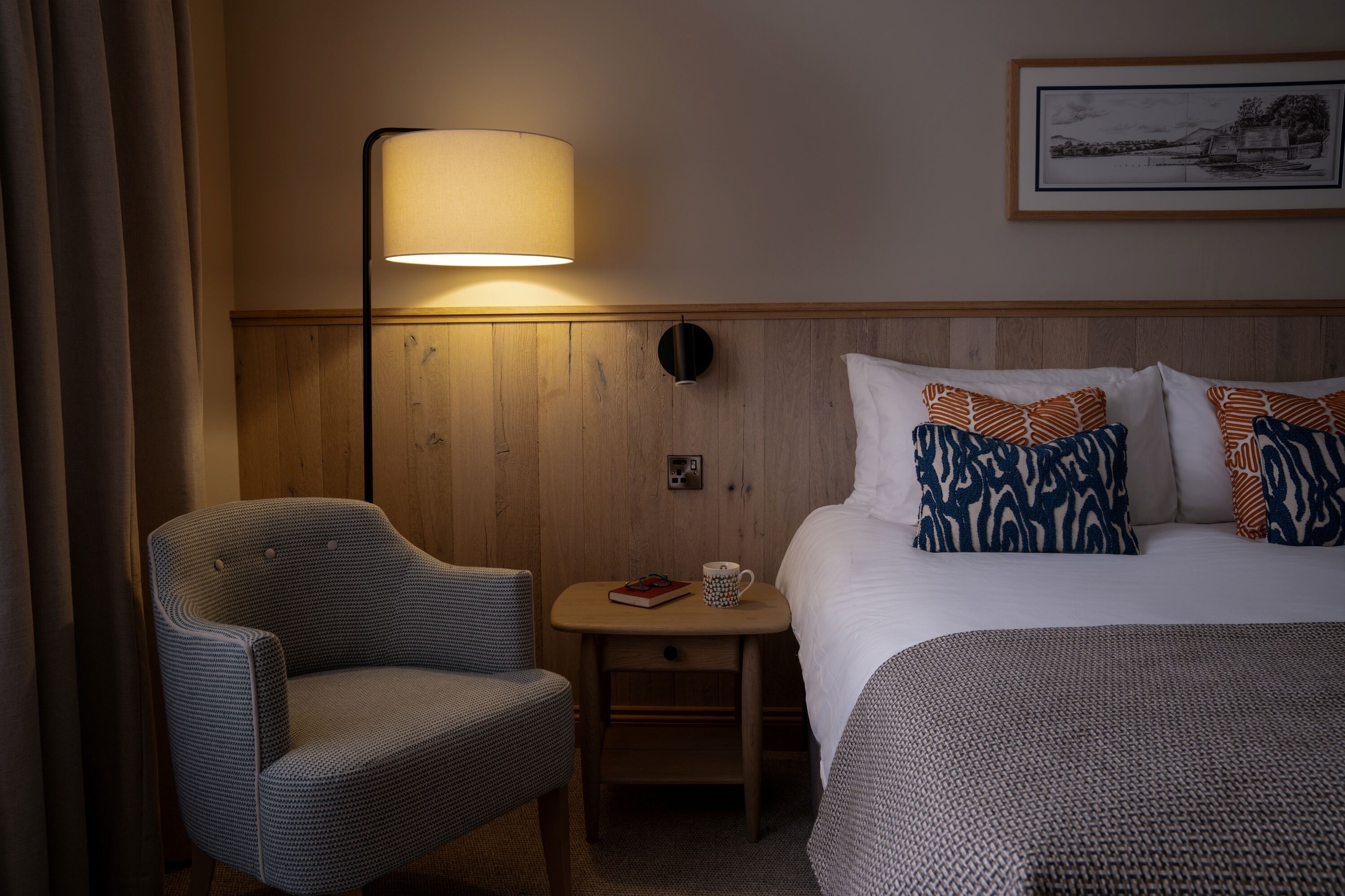 Habitación de hotel con una cama, una silla, una mesita de noche con lámpara y un cuadro en la pared.
