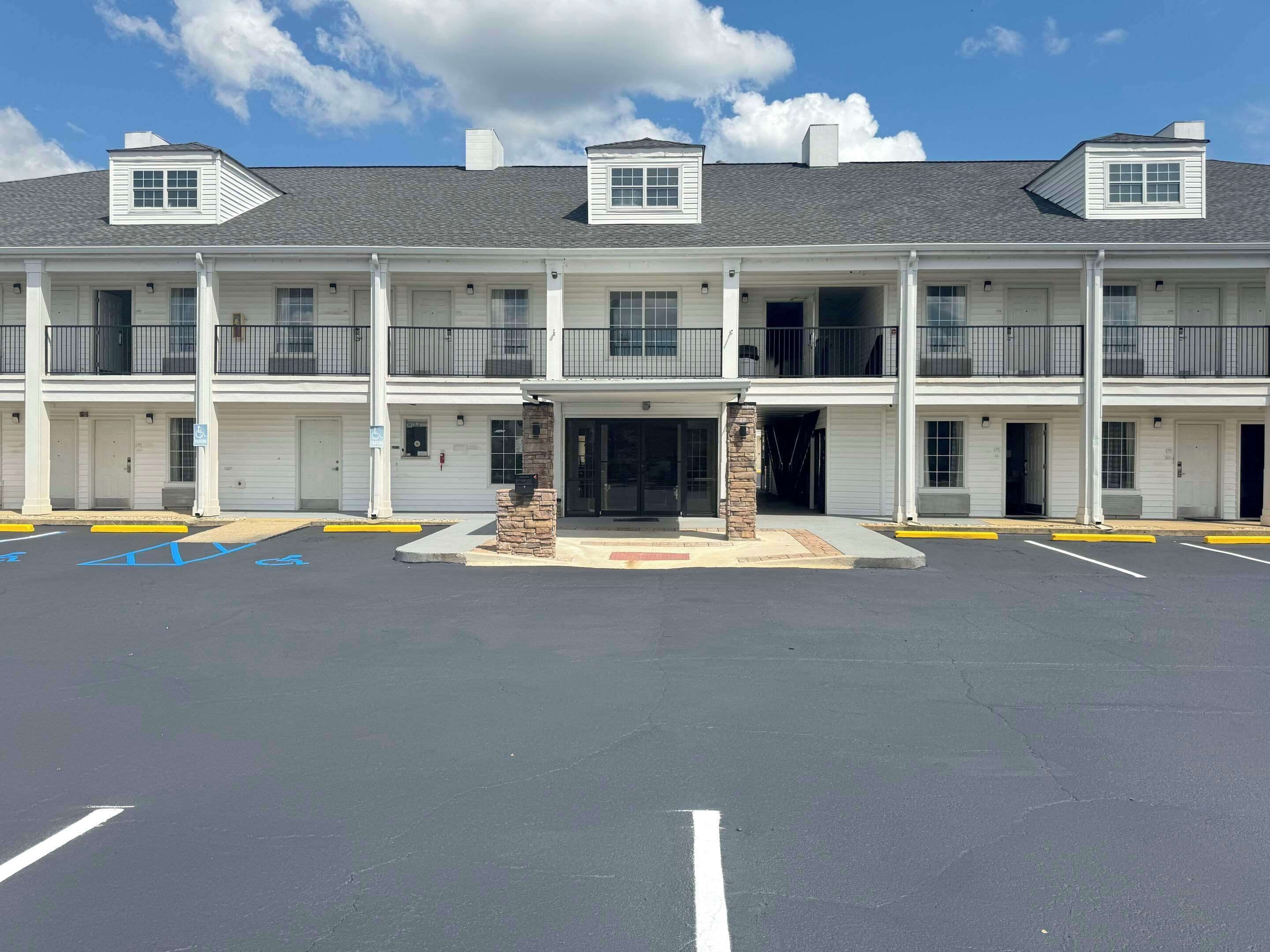 Un motel de varios pisos con entrada central, estacionamiento con espacios reservados para discapacitados y un cielo azul despejado.