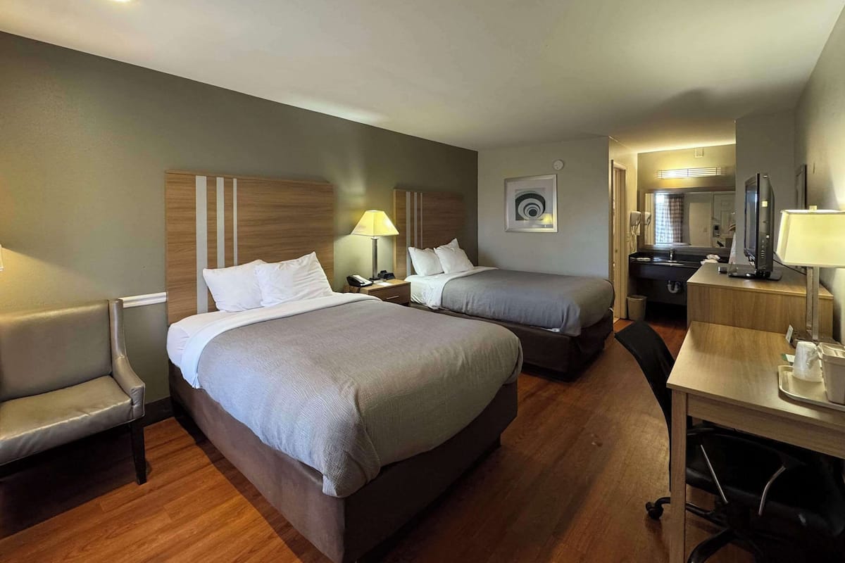 Habitación de hotel con dos camas, un escritorio, una silla, un televisor y un baño visible a través de una puerta abierta.