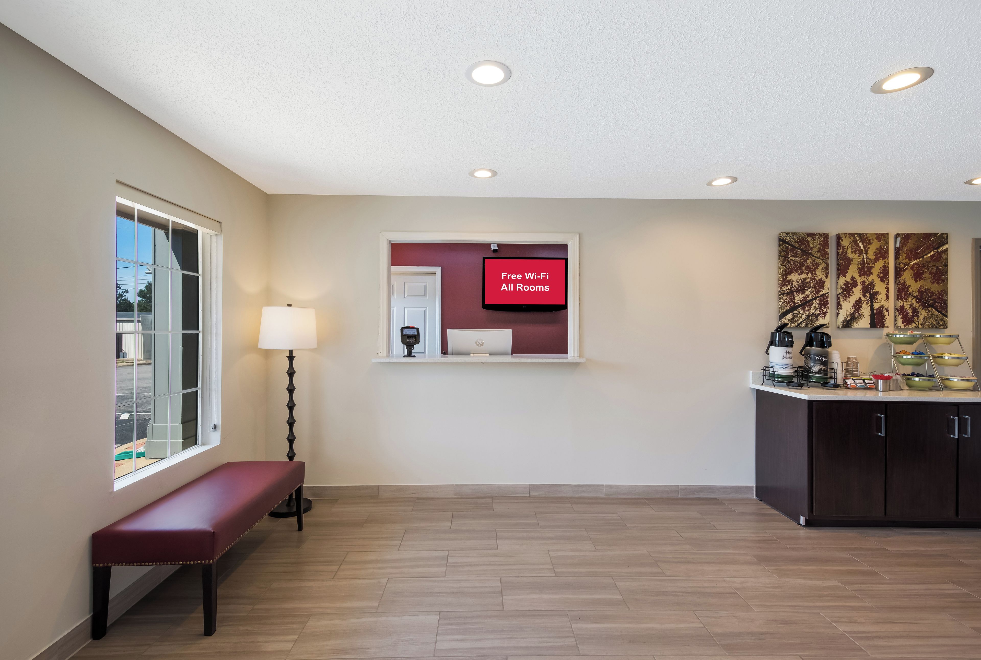 A hotel reception area with a bench, a wall-mounted TV displaying 'Free Wi-Fi All Rooms,' and a kitchen counter with various items.