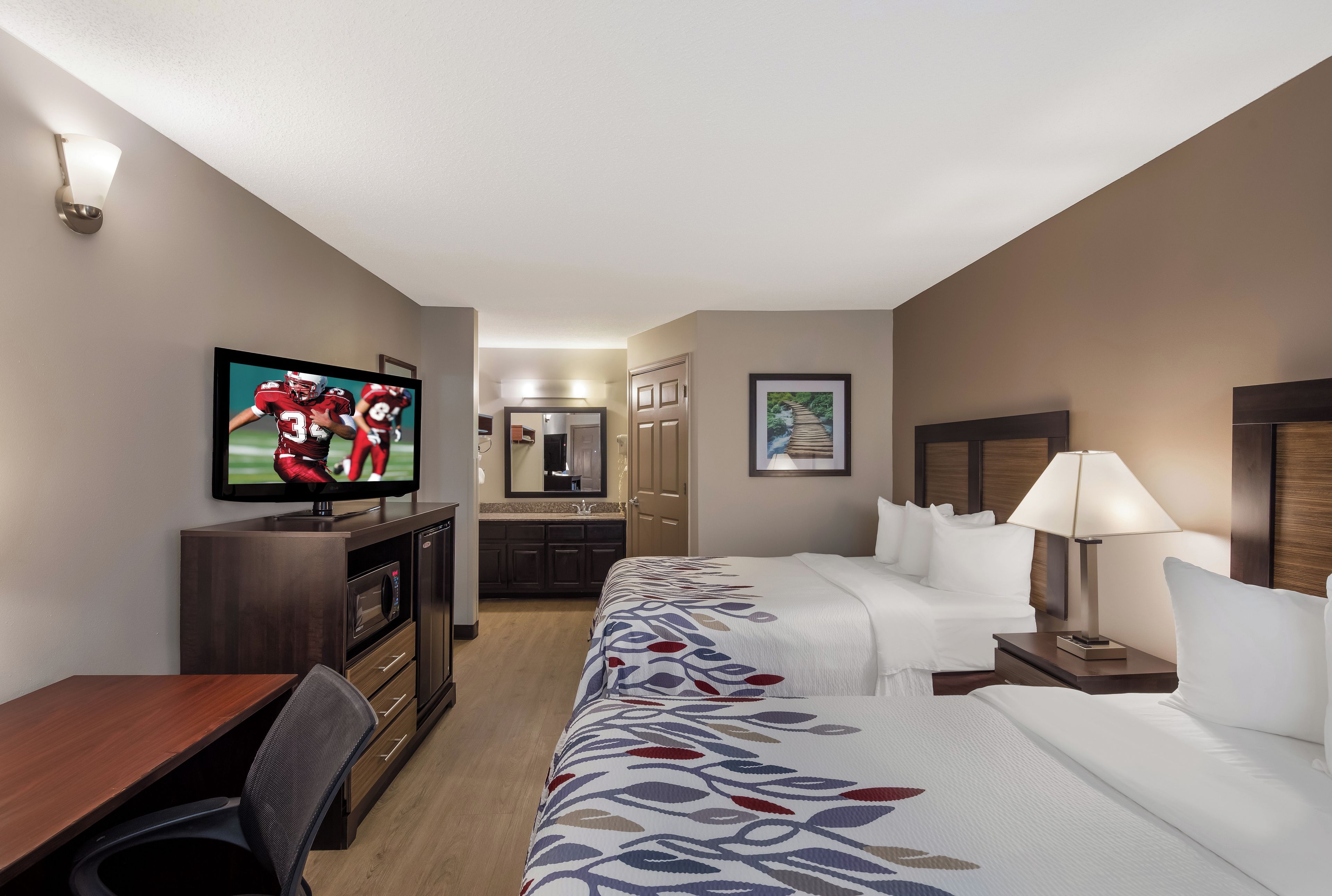 A hotel room with a TV, a desk, a bed, a nightstand, and a bathroom visible through an open door.