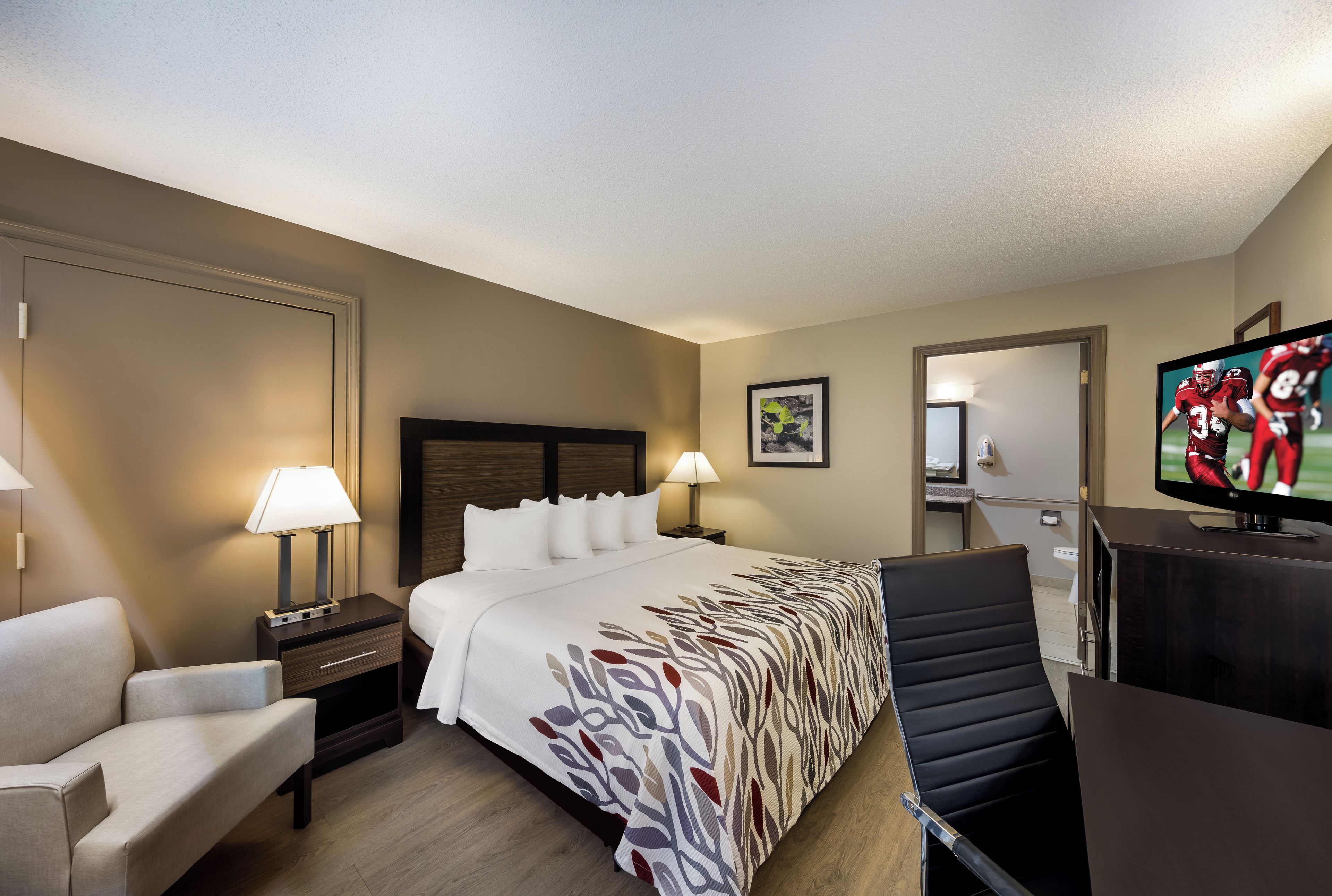 A hotel room with a bed, a desk, a chair, a television, and a bathroom visible through an open door.