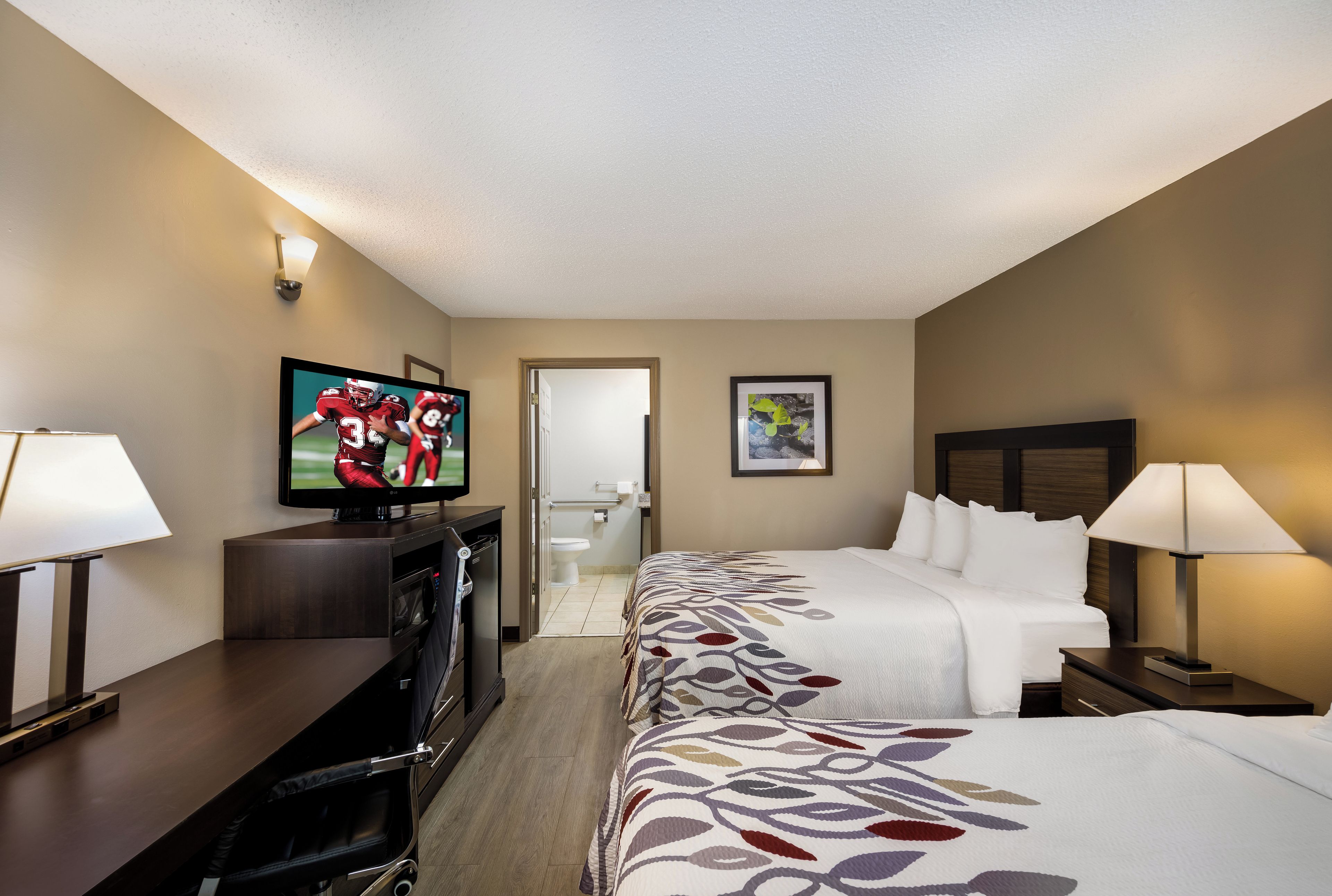 Ein Hotelzimmer mit Fernseher, Bett, Schreibtisch, Stuhl, Nachttisch und Lampe.