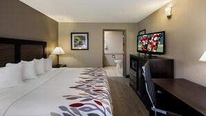 Habitación de hotel con cama, televisor, escritorio y un baño visible a través de una puerta abierta.